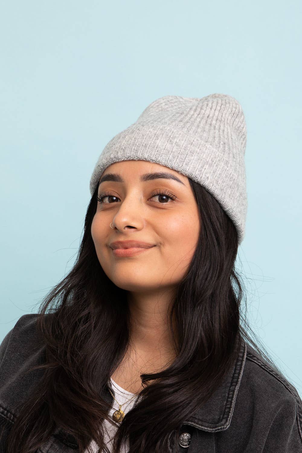 A young woman with long dark hair wears the Leto Accessories Versatile Cuffed Beanie—Cozy Rib Knit Hat in light gray, paired with a white top and denim jacket against a light blue background—the perfect fall accessory.