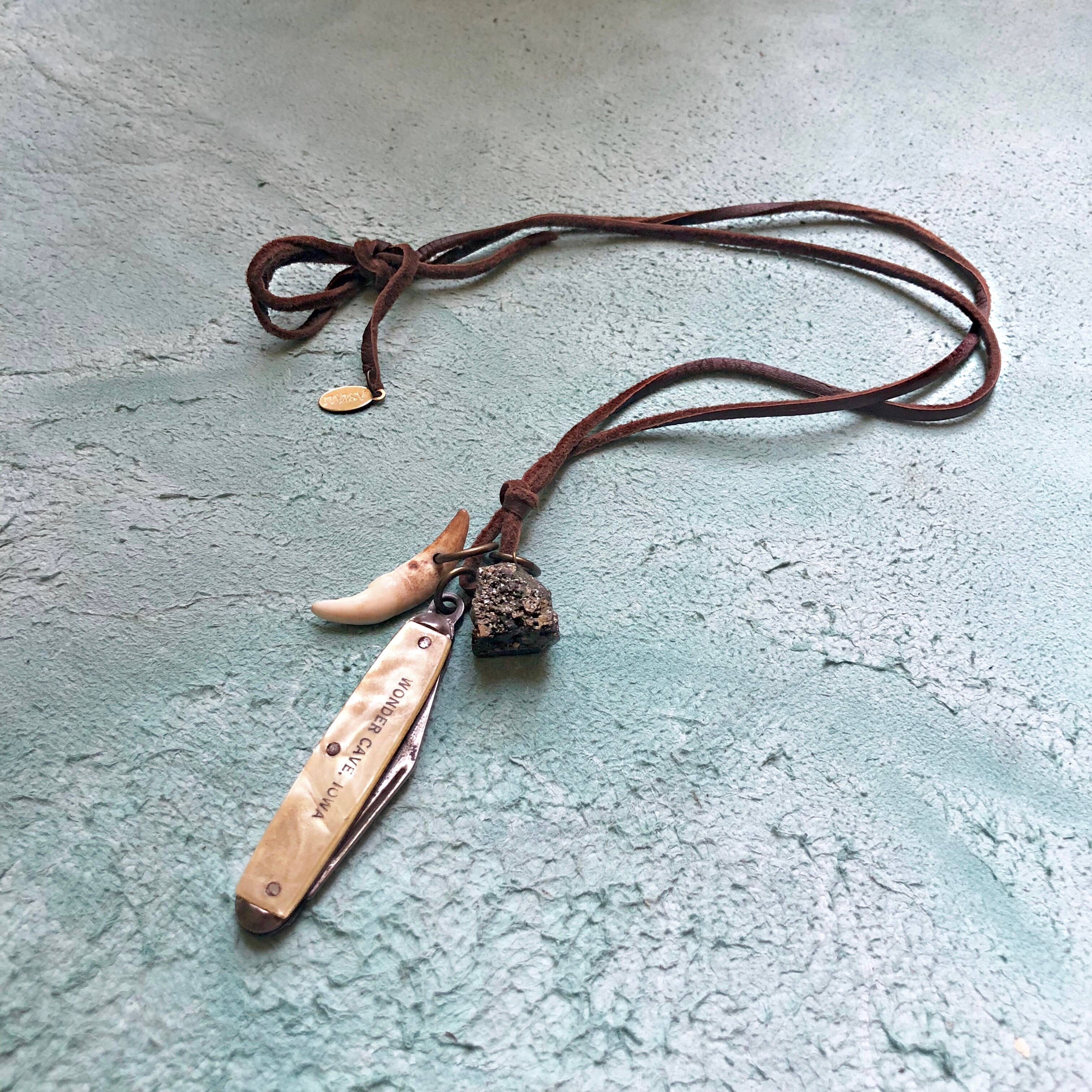The ASTALI Vintage Pocket Knife & Coyote Tooth Necklace features a pyrite stone and deerskin leather cord, displayed on a textured light blue surface.