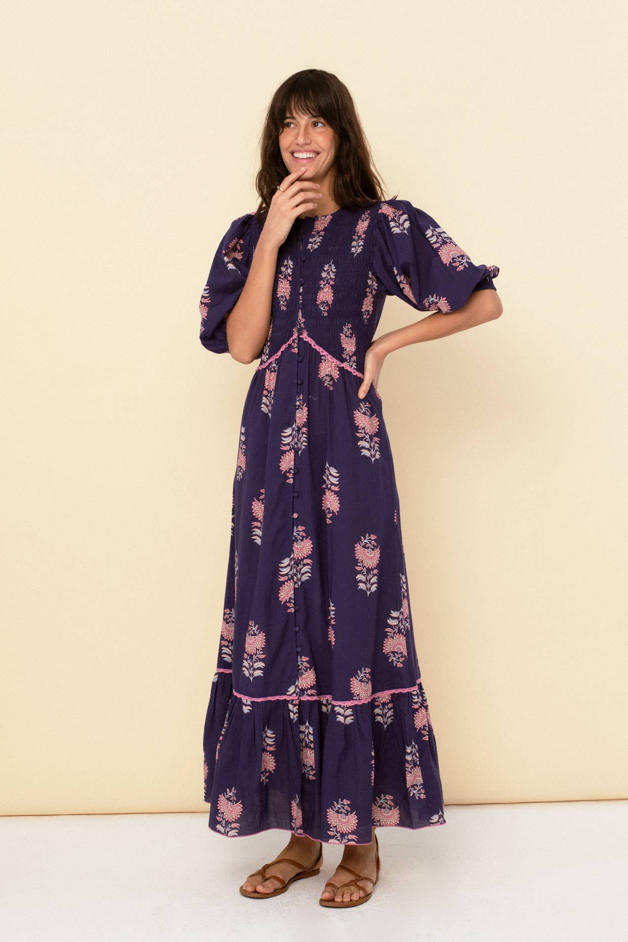 A woman stands against a beige background, smiling with one hand near her mouth. She wears the Magnolia dress blue by Sonmer Shop USA, featuring a romantic vintage style with puffed sleeves, paired with brown sandals.