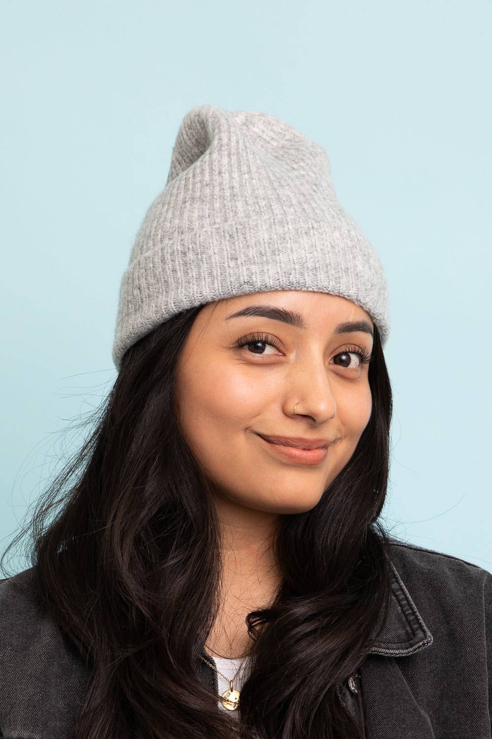 A woman with long dark hair wears the Versatile Cuffed Beanie – Cozy Rib Knit Hat by Leto Accessories in light gray with a dark jacket, smiling softly against a light blue background—the ideal fall accessory.
