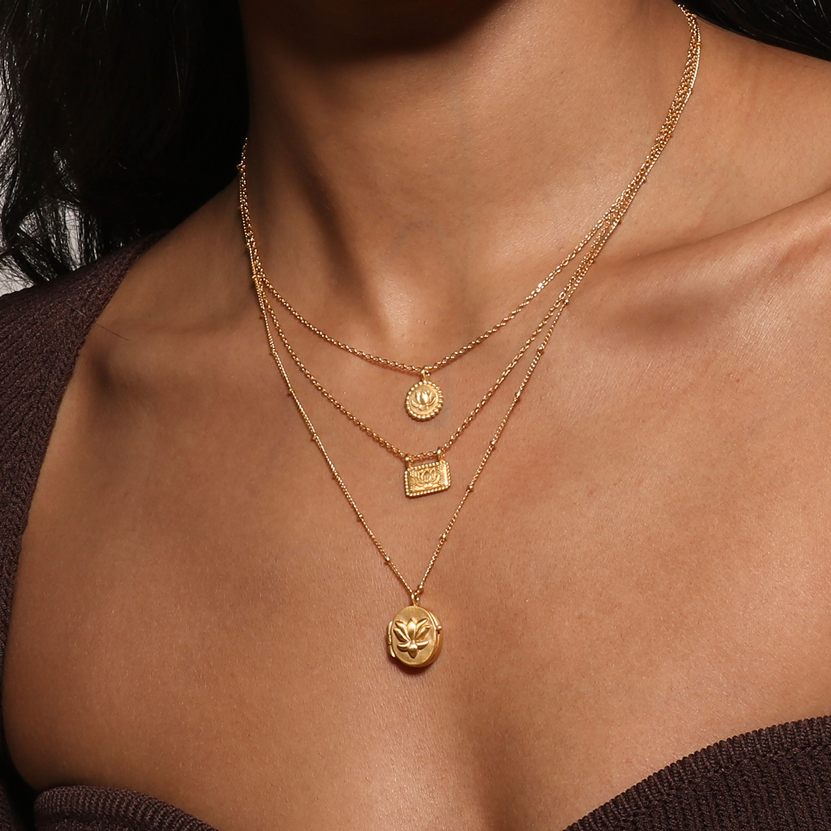 A woman wears the Satya Jewelry 16” Lotus Plate Necklace, featuring three layered gold-plated chains with round and rectangular pendants, including a lotus symbolizing new beginnings, paired with an off-shoulder brown top.