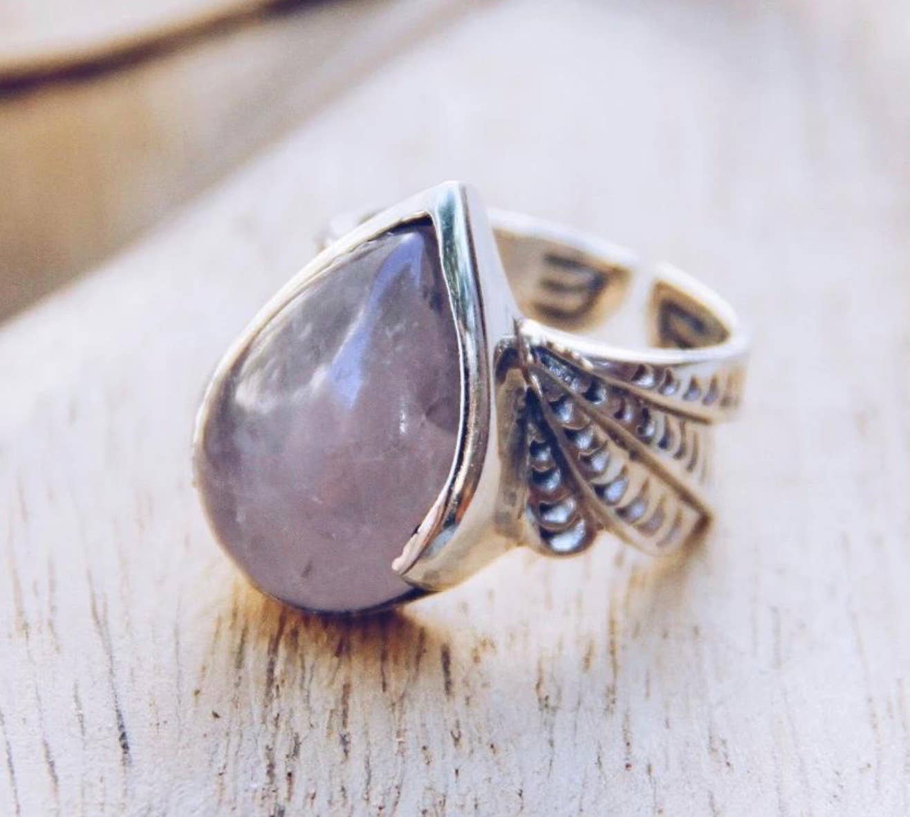The bahgsu jewels Winged Ring features a silver ornate band with a large teardrop-shaped pale purple gemstone at the center, displayed on a light wooden surface.