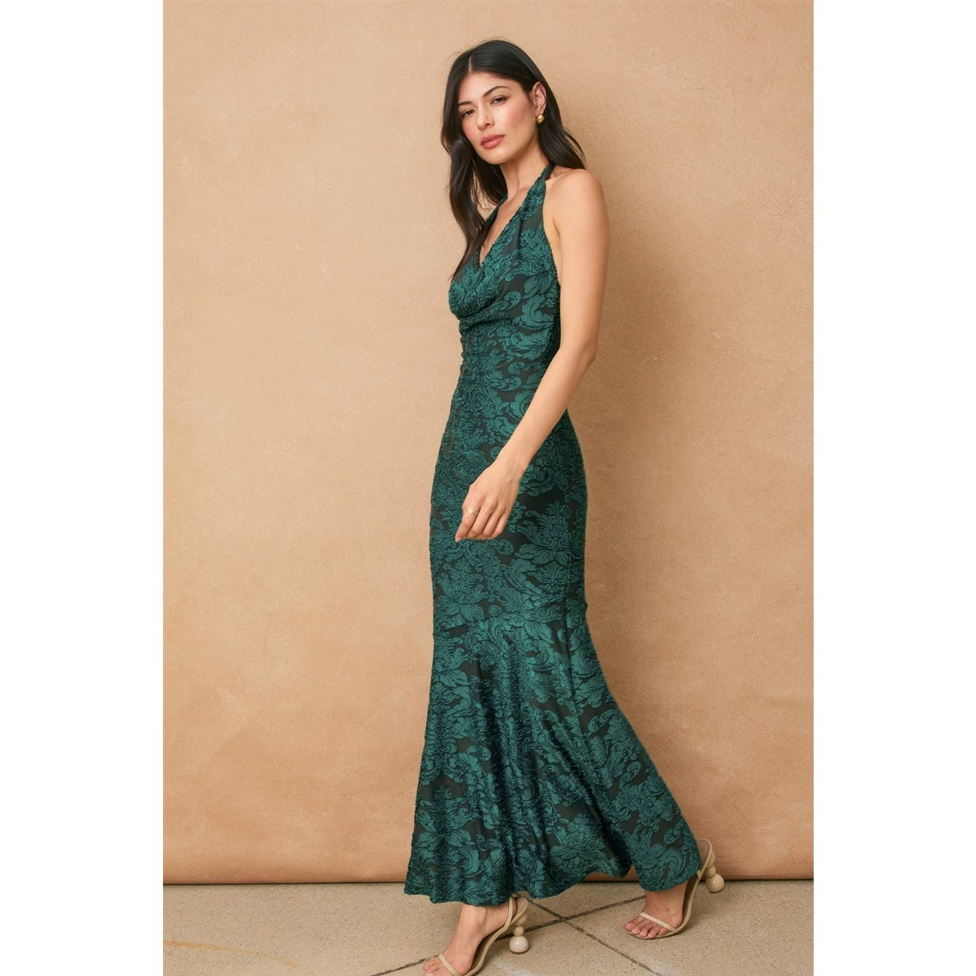 A woman in profile models the Dress Forum Fame Draped Halter Neck Maxi Dress, a floor-length, sleeveless emerald green gown with a floral pattern. She has long dark hair and neutral makeup, posed against a plain beige background.