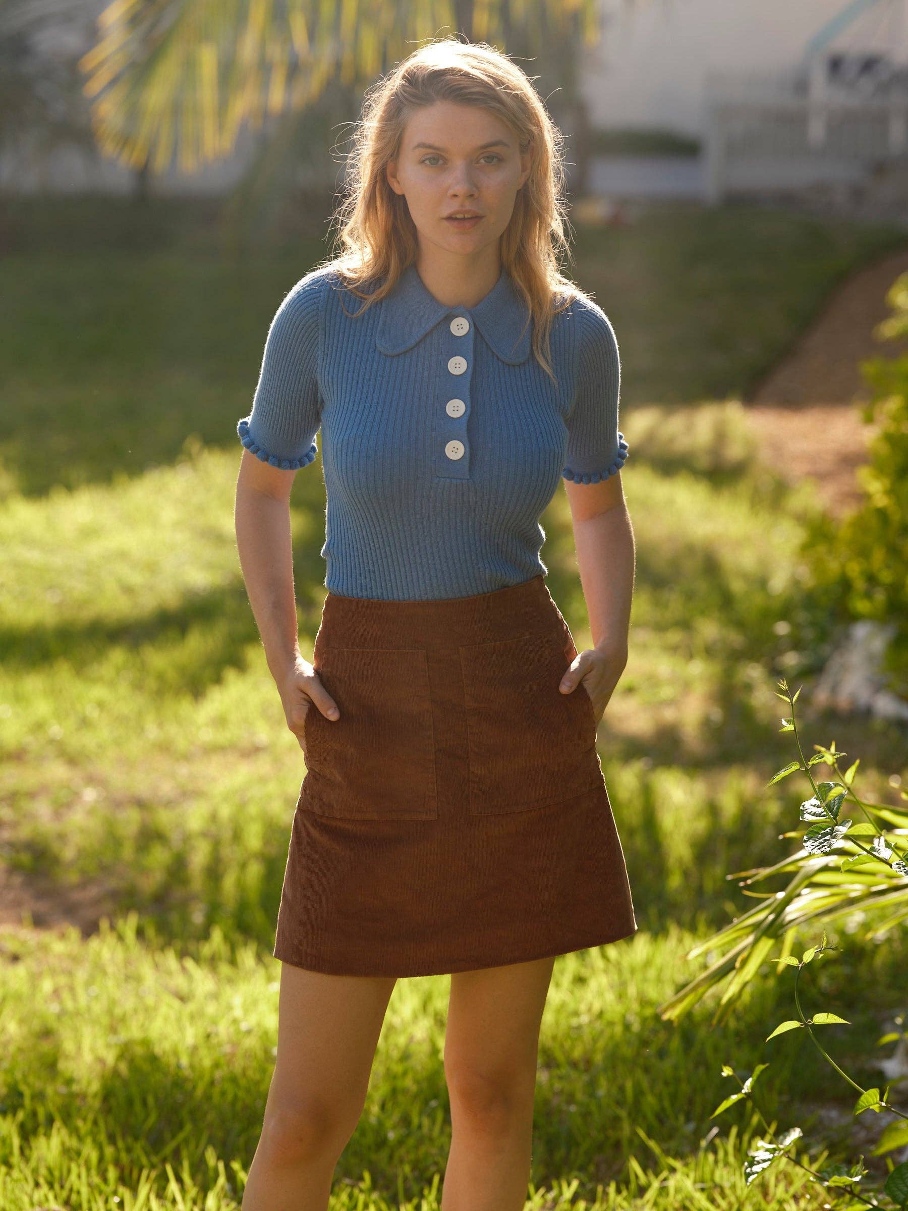 A woman stands on grass wearing the Mollusk Belle Tee, a vintage-inspired blue short-sleeve knit top with white buttons, paired with a brown skirt. Sunlight highlights her hair amid lush greenery in the background.