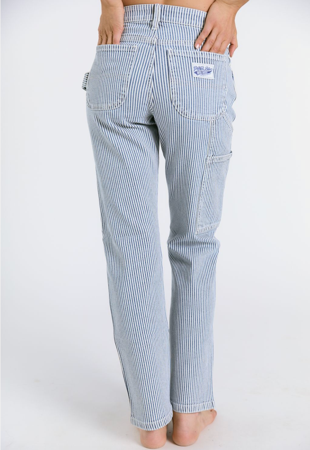 A person stands barefoot on a white background, wearing Porter Blue Apparels PEYTON PAINTER IN PACIFIC WASH jeans. The photo shows the back view with hands on the waistband and visible back pockets.