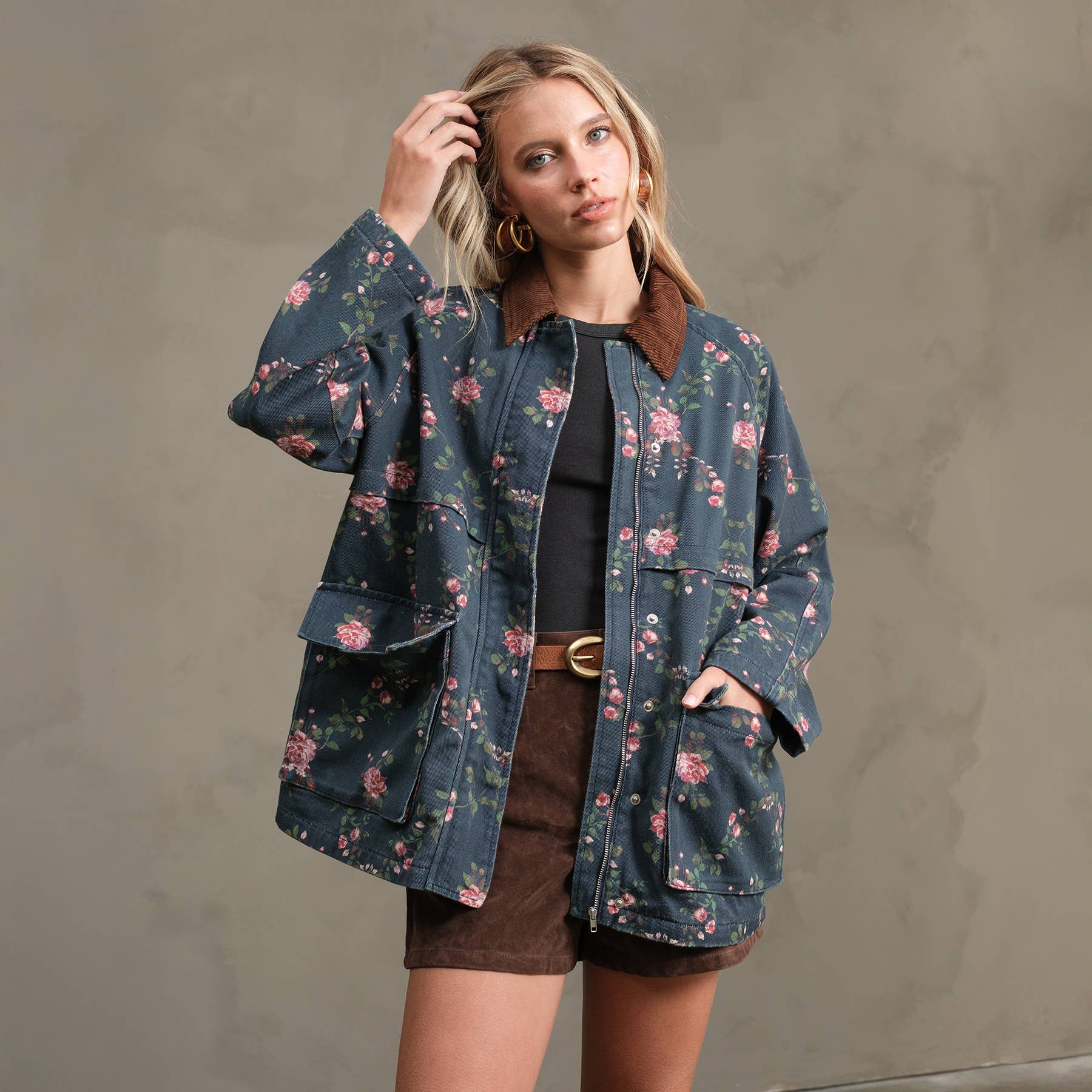 A woman with blonde hair poses against a plain background, wearing the RAISONNEL Floral Printed Utility Jacket (RJ1073) with a brown collar, paired with a black top and brown skirt, her hand resting on her head as she looks at the camera.