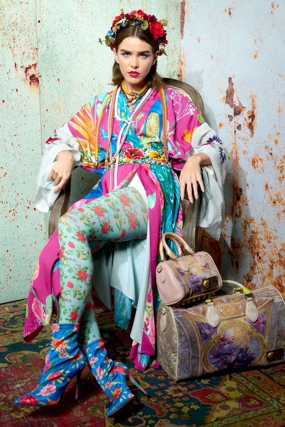 A woman sits on a chair against a rustic wall, wearing the Aratta Marie Antoinette Kimono with embroidered floral sleeves and a flower crown. She poses seriously, with two ornate handbags by her side.