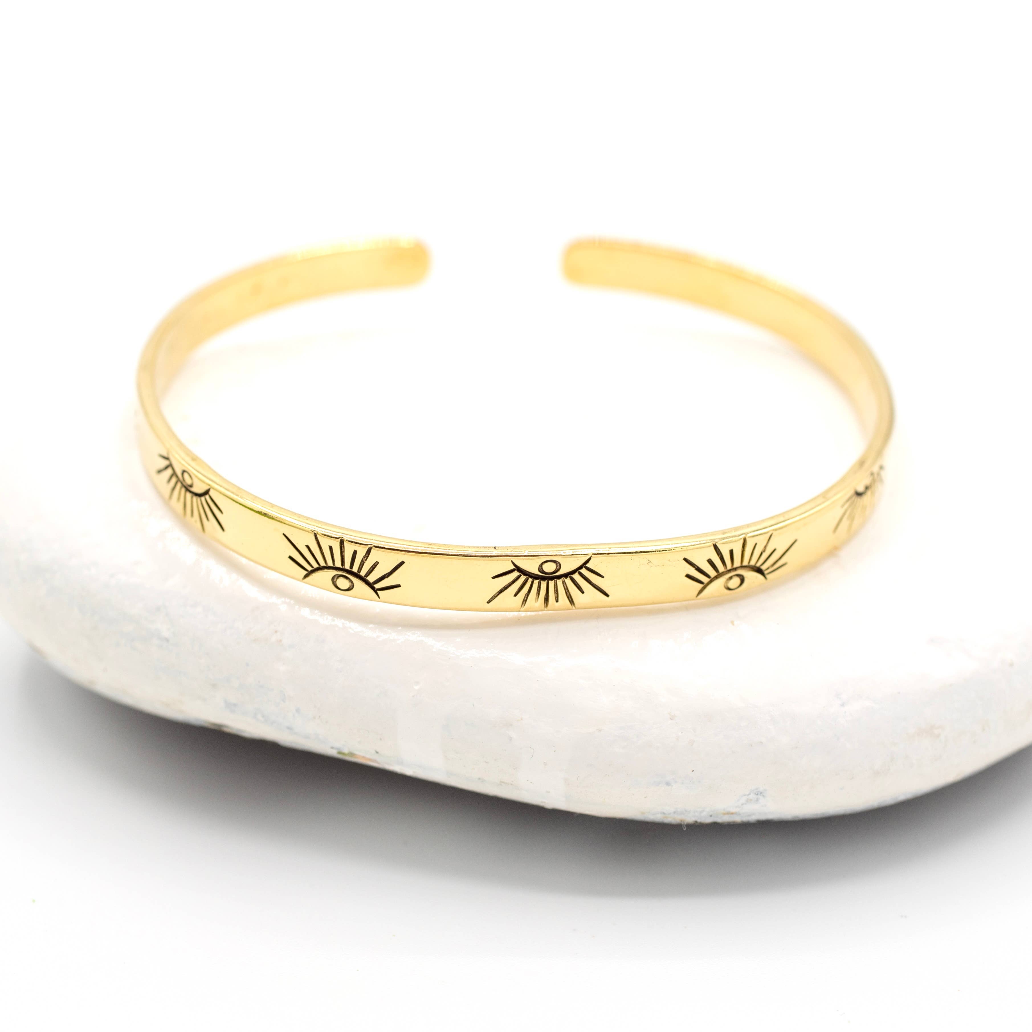 The Baizaar Brass Eye Bangle, featuring a repeating eye pattern, is displayed on a white stone background—ideal for fans of boho and unique handmade jewelry.