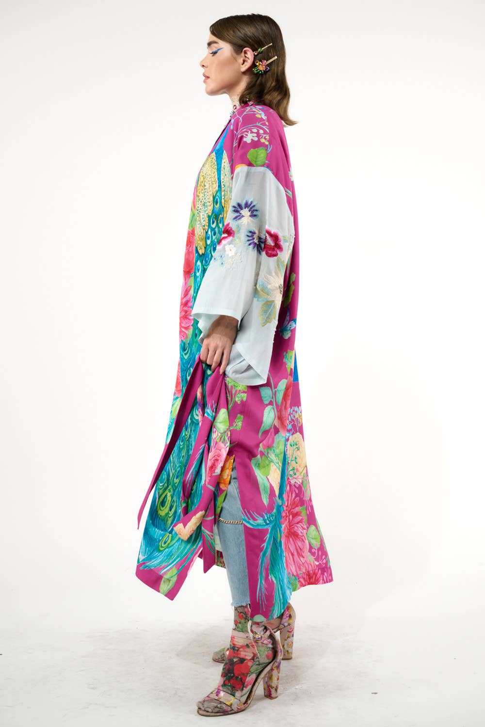 A person stands in profile wearing the vibrant Marie Antoinette Kimono by Aratta, featuring embroidered floral sleeves, styled with light blue jeans and floral-patterned boots, making a bold statement against a white background.