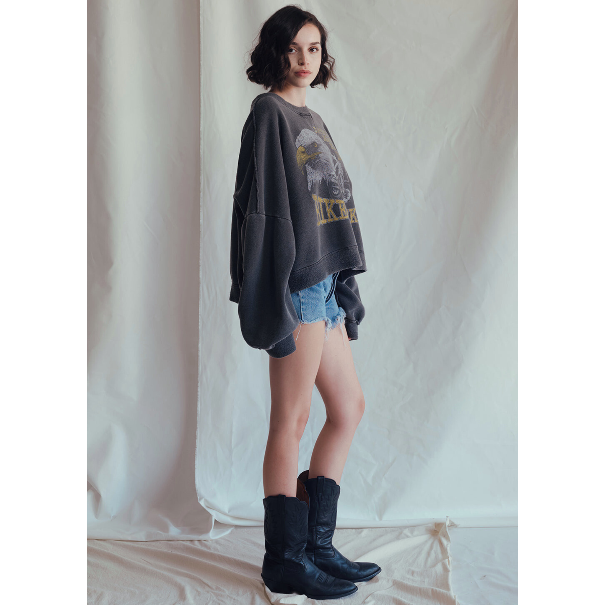 A young woman with short dark hair wears the People Of Leisure Daytona Sweatshirt featuring a vintage eagle graphic, paired with denim shorts and black cowboy boots, standing sideways against a light fabric backdrop.