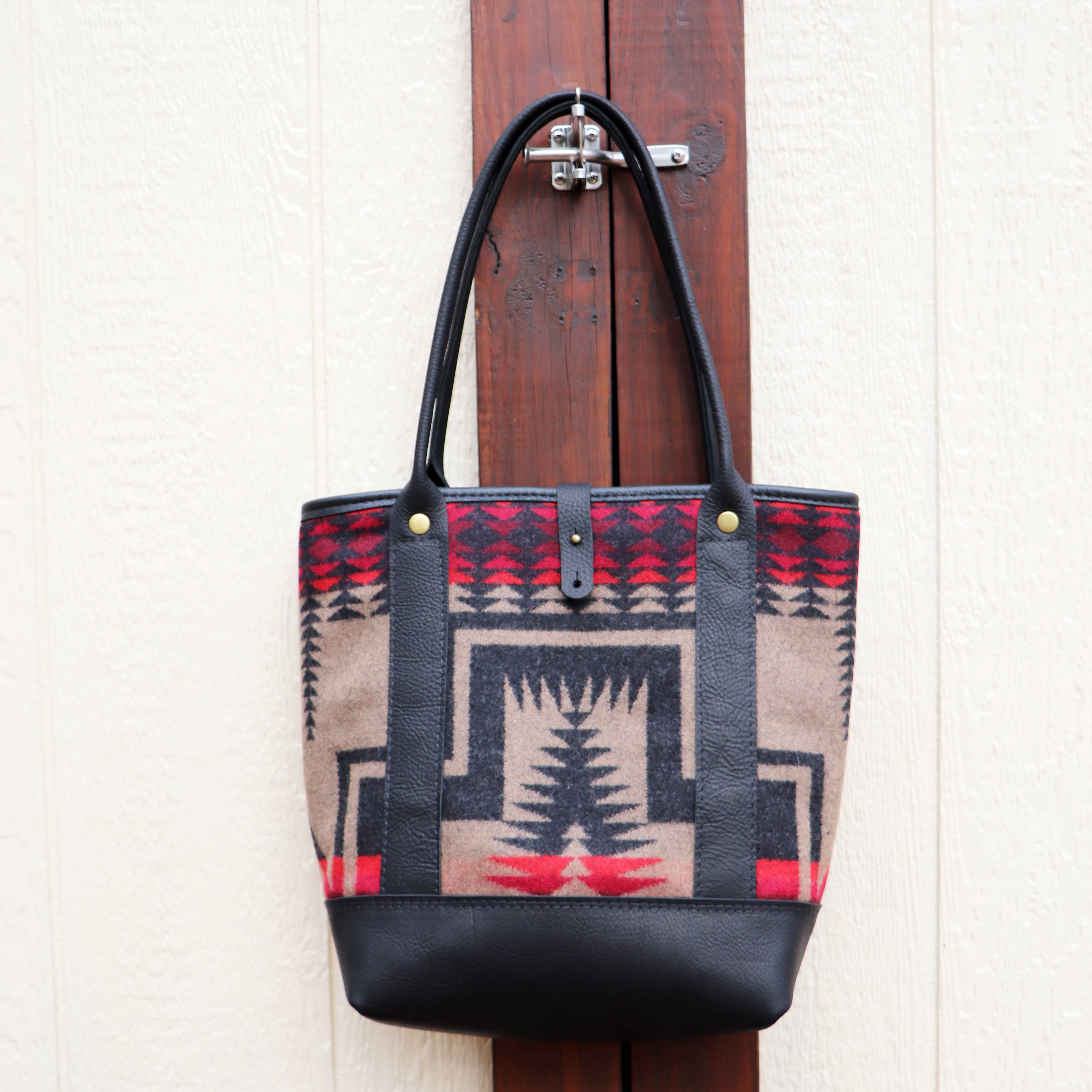 The Hawks & Doves Pearl Wool and Leather Tote, with black full grain leather handles and base, features a geometric Native American-inspired pattern in black, red, and beige and hangs on a hook by a cream wall with wood trim.