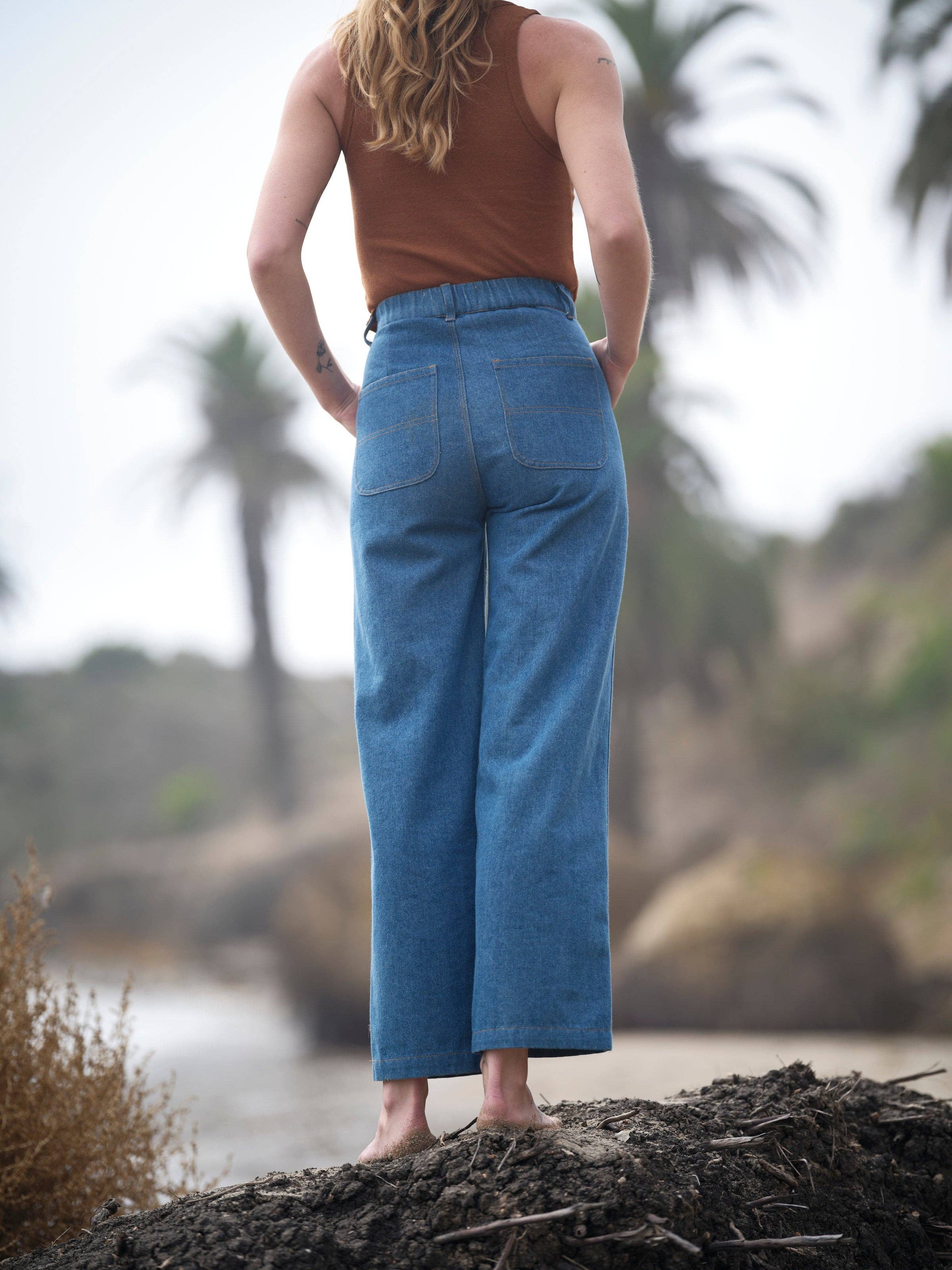 A person wears Mollusk Painter Pants in Indigo and a brown sleeveless top, standing barefoot on sand outdoors with palm trees and a blurred natural background.