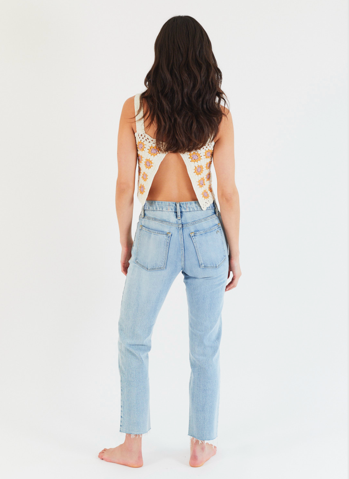 A woman with long wavy brown hair stands barefoot, facing away, wearing Porter Blue Apparels REBEL STRAIGHT IN EDEN WASH high-rise jeans and a sleeveless white top with orange floral patterns, set against a plain white background.