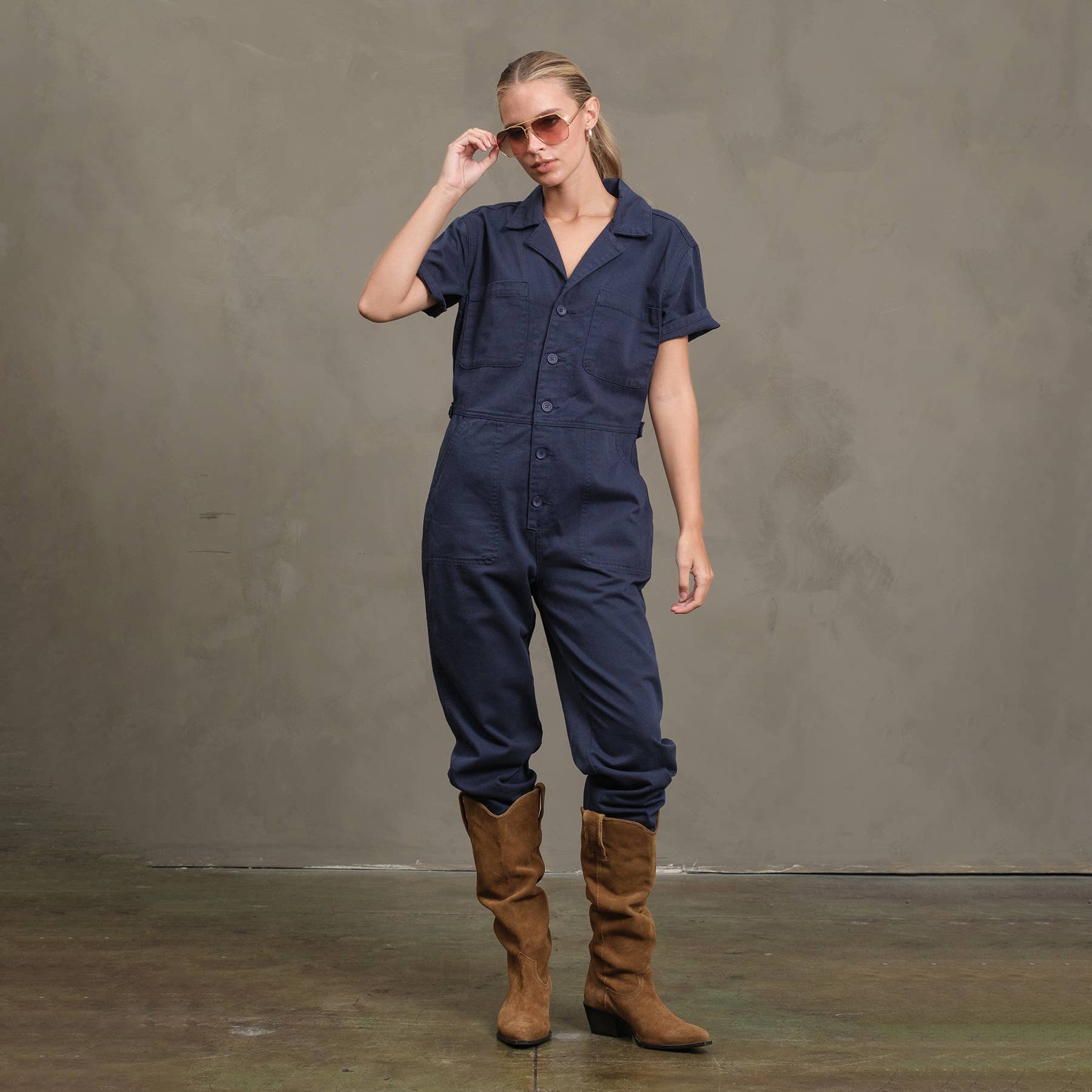 Standing against a plain gray wall, a person wears the RAISONNEL Field Jumpsuit - RJP1001 in navy blue with rolled sleeves, paired with brown suede boots. Their long hair is pulled back as they adjust sunglasses with one hand.