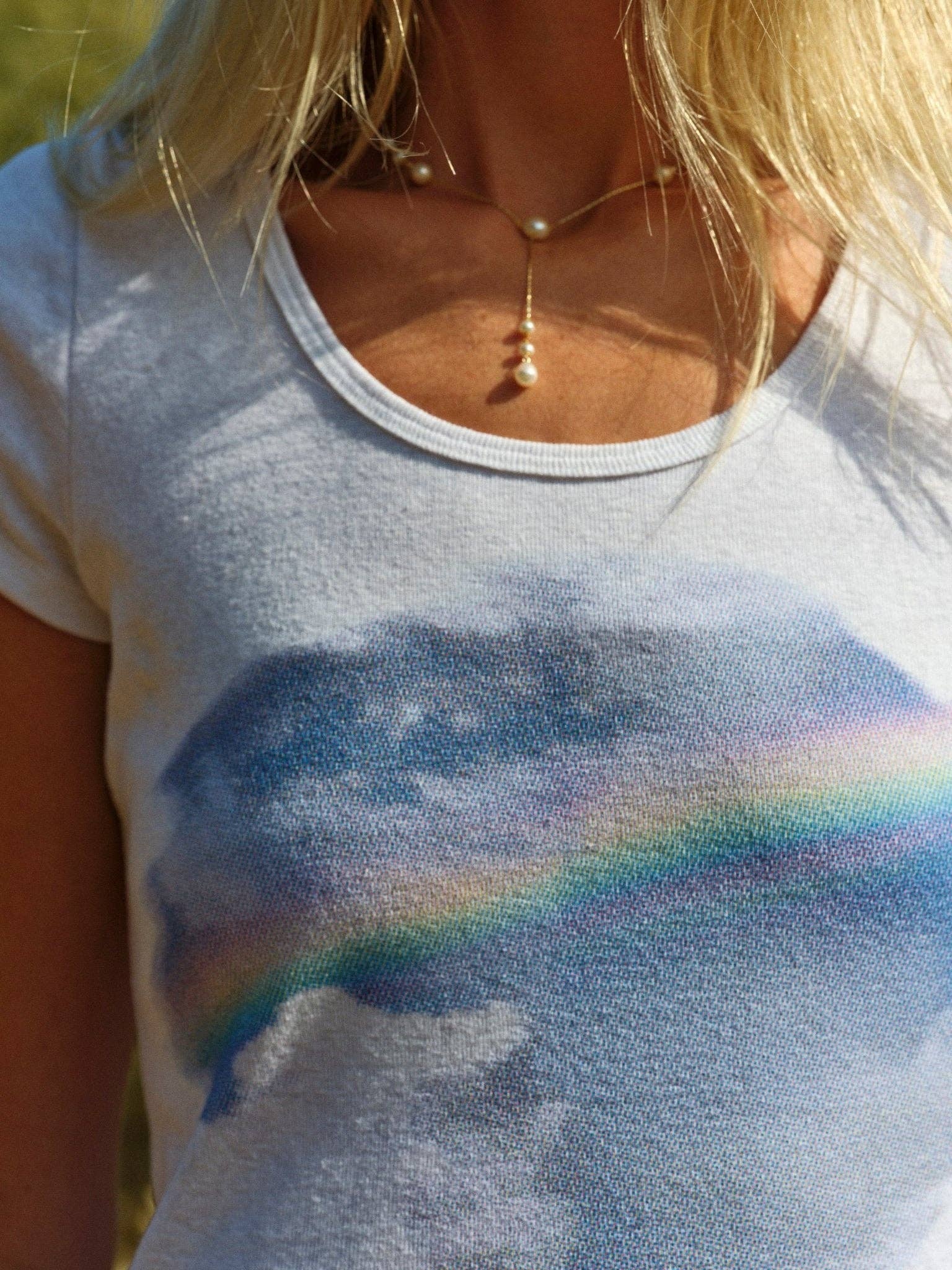 A close-up shows a person in the Mollusk CloudBreak Tee, a white hemp cotton shirt with rainbow and clouds graphic, styled with a pearl necklace. The look channels relaxed California surf vibes, with long blonde hair partially visible.