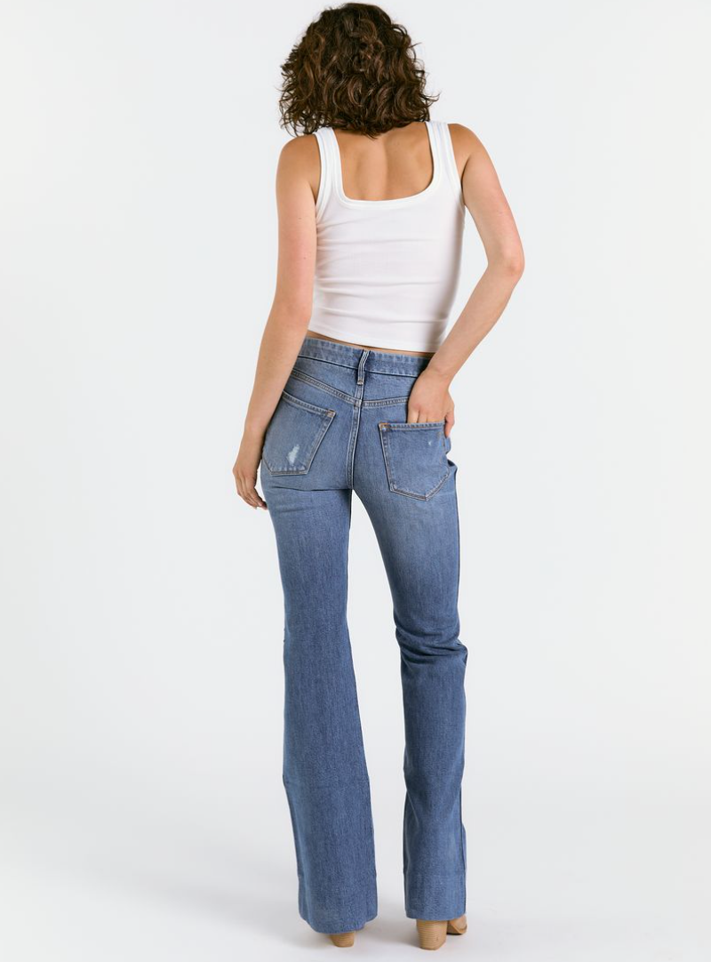 A person with curly hair, wearing a white tank top and Porter Blue Apparel’s WANDERER 5 POCKET FLARE IN MABEL WASH, stands facing away with one hand in their back pocket against a plain white background.