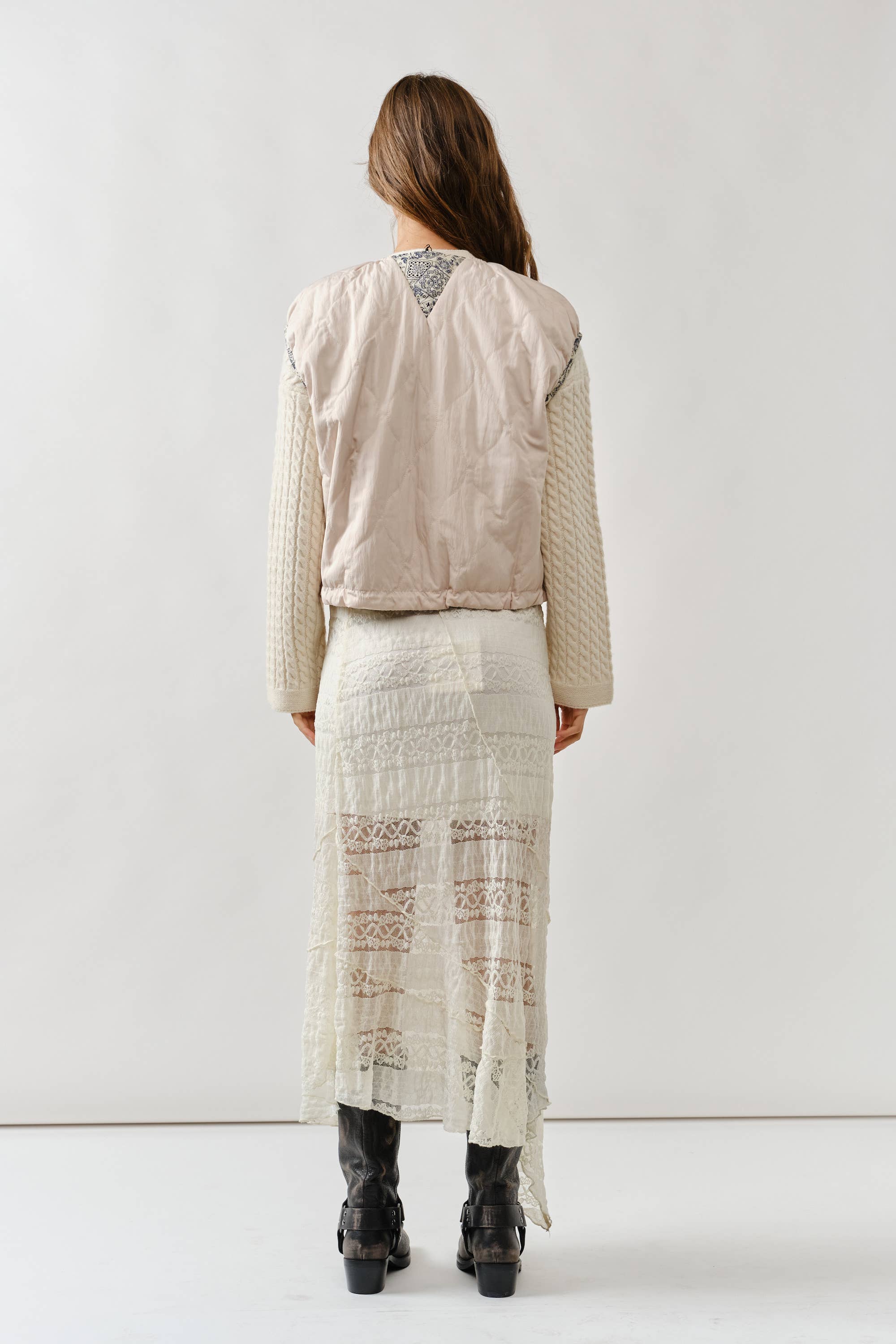 A woman with long brown hair faces away, wearing the Aaron & Amber Contrast Detail Lightweight Vest layered over a cream lace skirt and paired with black heeled boots, set against a plain white background.