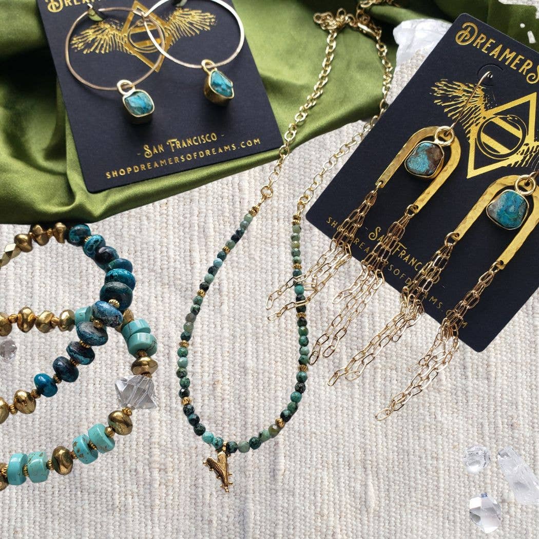 A display of handmade bohemian jewelry from Dreamers of Dreams features the Chrysocolla Beaded Heart Charm Necklace and gold earrings, elegantly arranged on a neutral fabric background.
