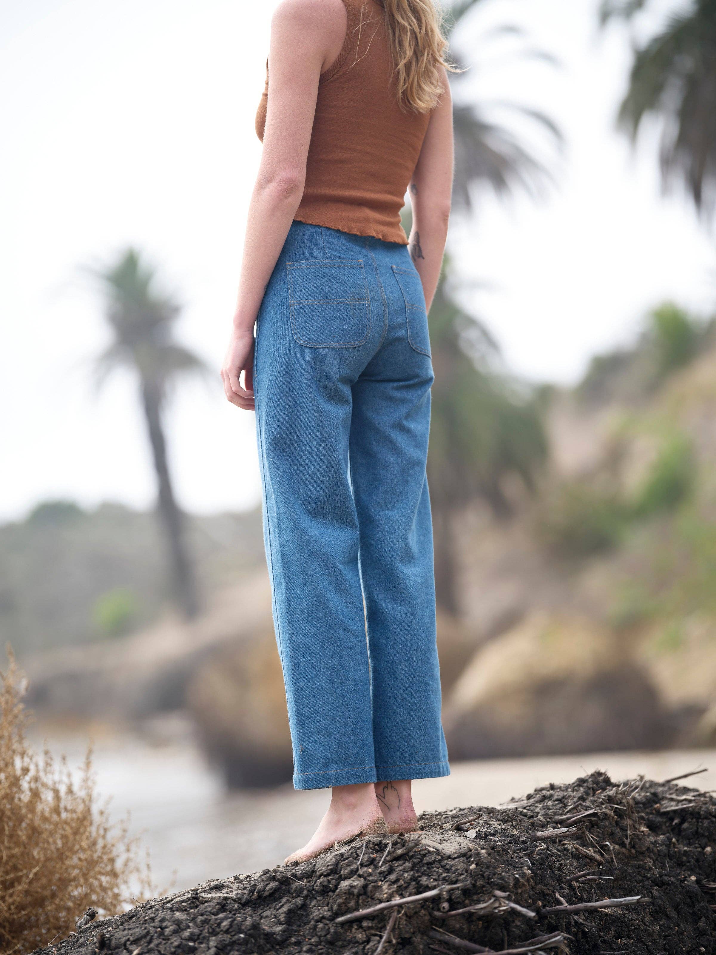 A woman with long hair stands barefoot on a dirt mound, wearing Mollusk Painter Pants in Indigo and a brown sleeveless top, with palm trees and a blurred natural background behind her.