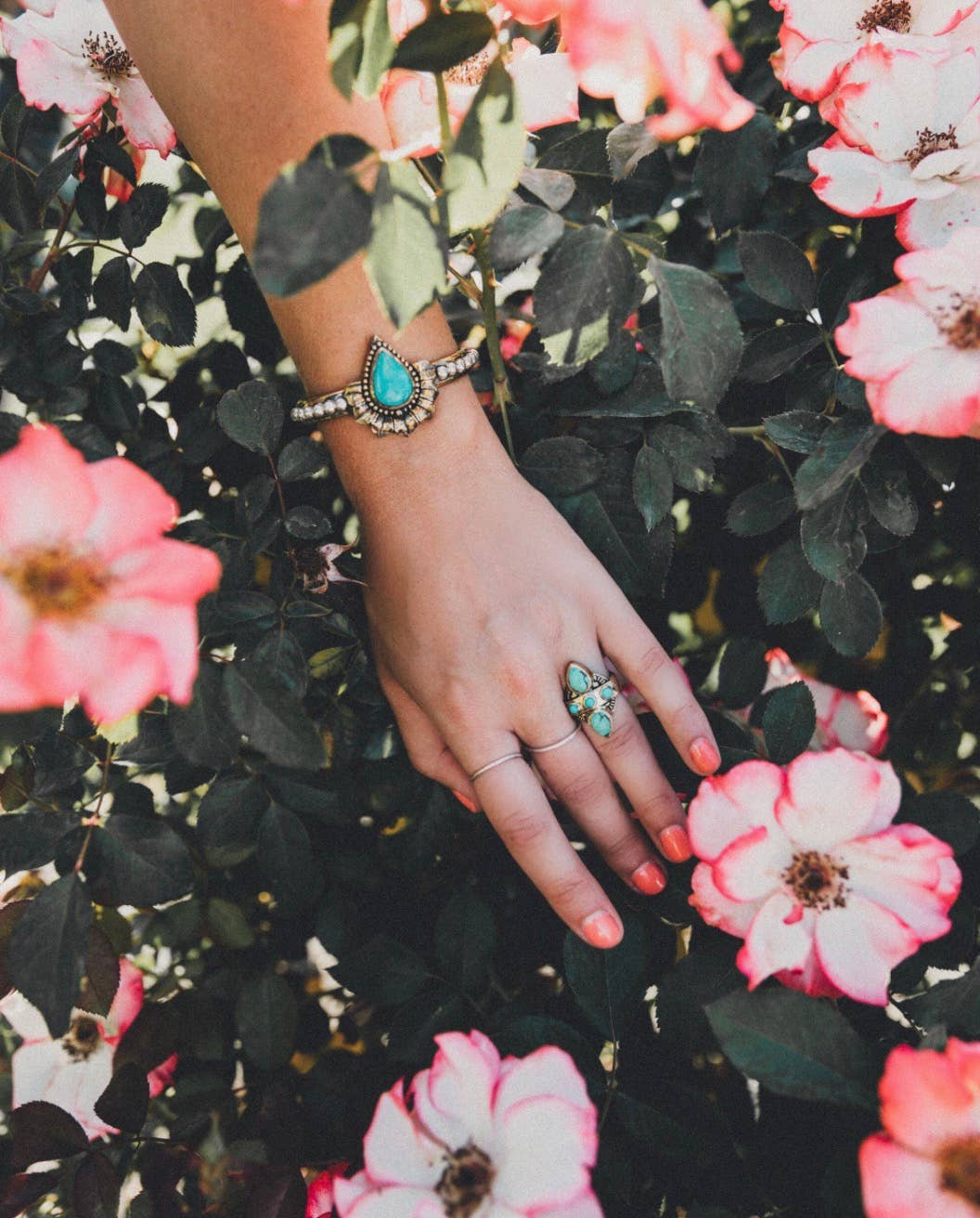 A hand with pink nails, adorned with the bahgsu jewels Truth Seeker Ring and a turquoise bracelet, rests among blooming pink and white flowers—a peaceful setting for jewelry lovers.