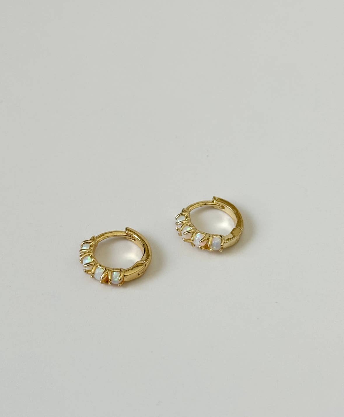 The Sonata Opal Hoops by Tramps+Thieves are miniature gold-plated huggie earrings, each accented with five iridescent opal stones along the front, shown against a plain off-white background.