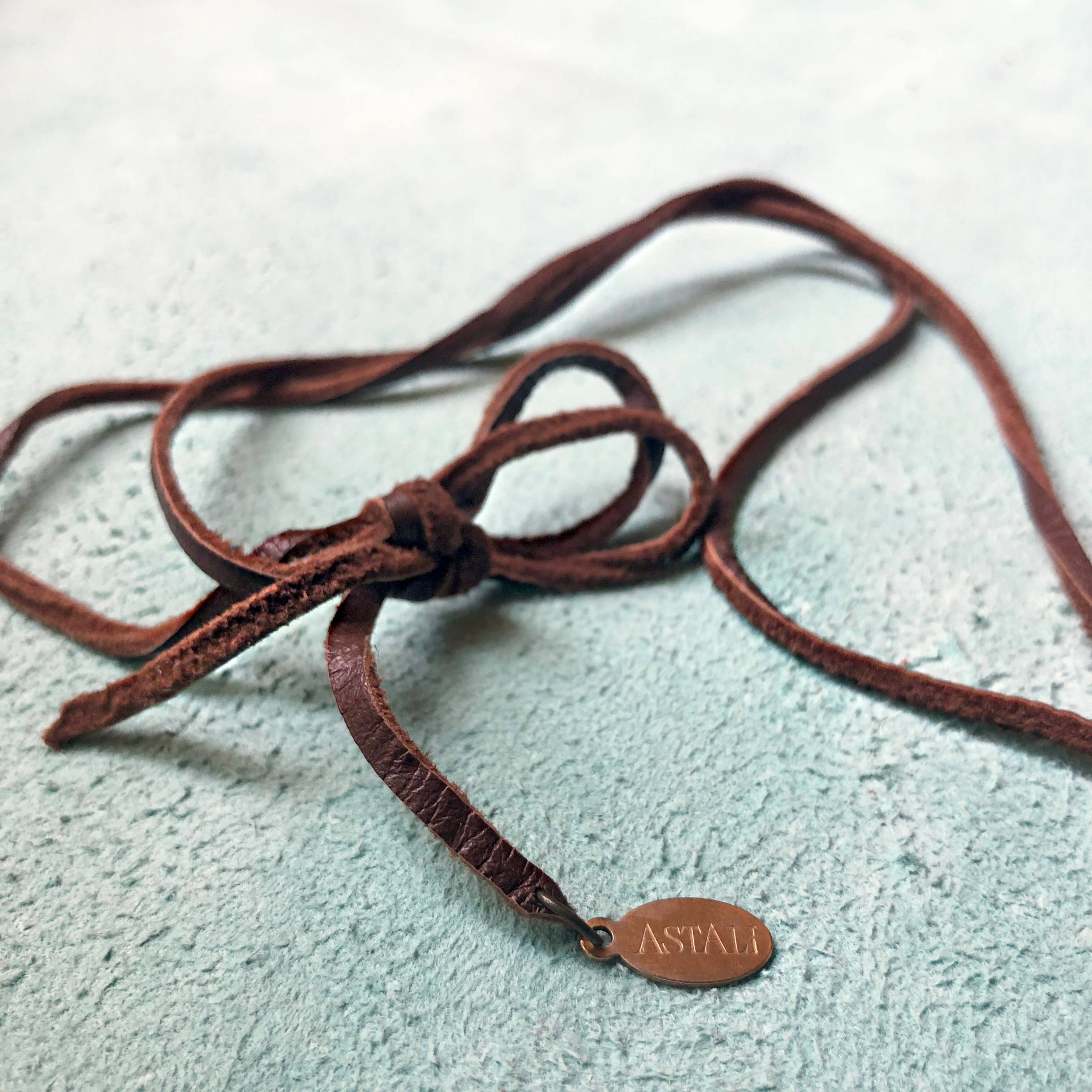 A deerskin leather cord tied in a bow rests on textured blue, beside an oval ASTALI tag, capturing the rustic charm of the Vintage Pocket Knife & Coyote Tooth Necklace – Pyrite & Deerskin Leather by ASTALI.
