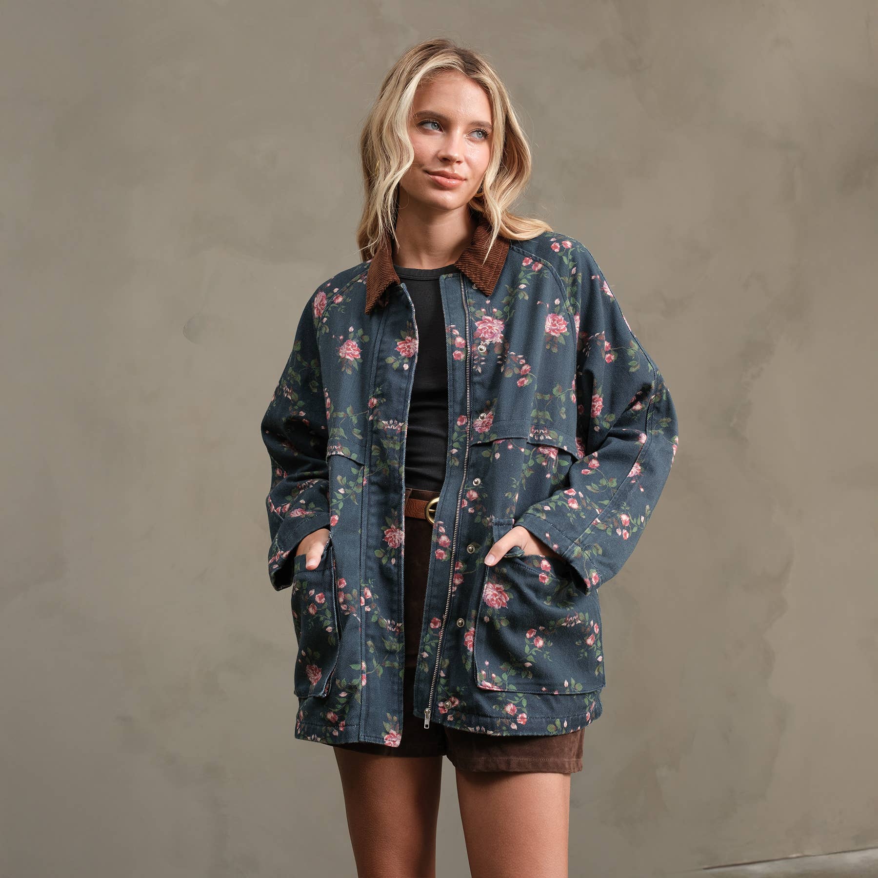 A woman wearing the RAISONNEL Floral Printed Utility Jacket (RJ1073) with brown trim, a black top, and brown shorts stands against a neutral background, hands in her jacket pockets and smiling softly as she looks to the side.