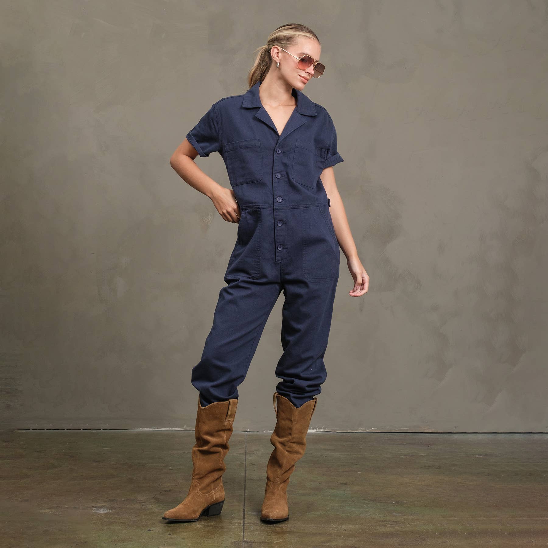 A person poses with one hand on their hip, wearing the RAISONNEL Field Jumpsuit - RJP1001 in navy blue, tan knee-high boots, and sunglasses on a concrete floor against a plain gray wall.