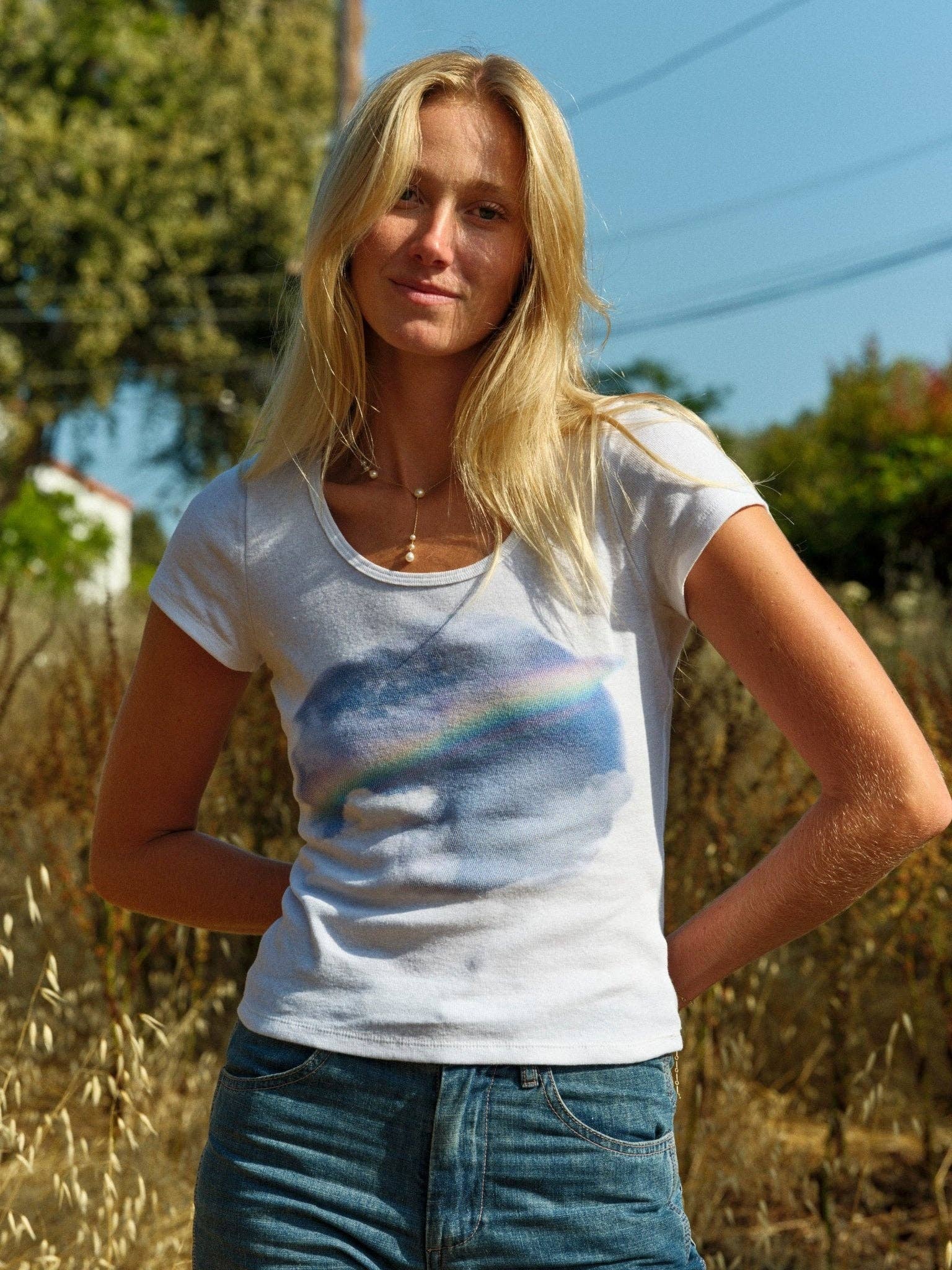 A blonde woman stands in tall grass outdoors, wearing the Mollusk CloudBreak Tee—a white tee with a pastel rainbow design—evoking California surf vibes on a sunny day with blue sky and trees behind her.
