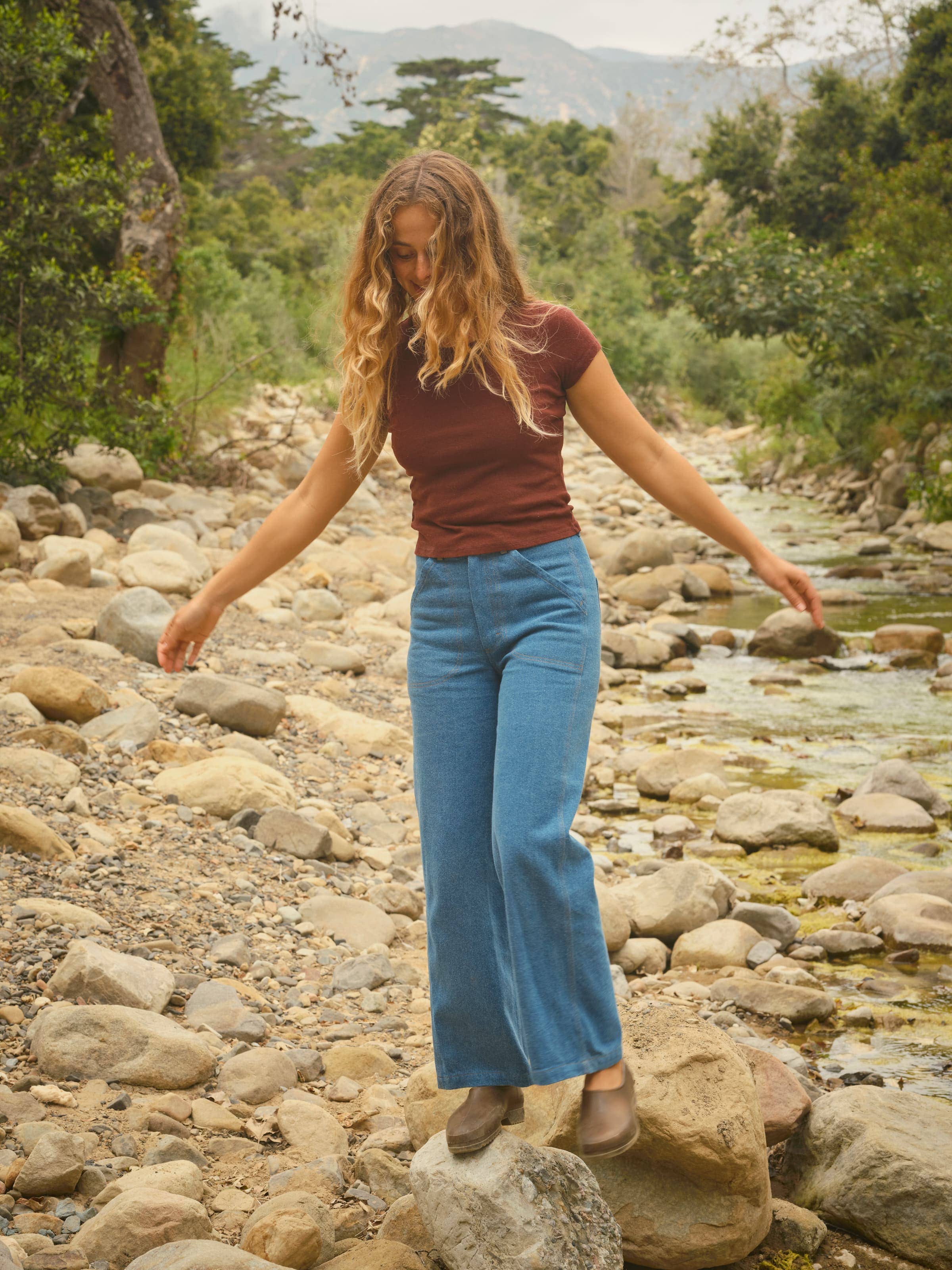 A woman with long wavy hair, in a maroon shirt and Mollusk Painter Pants in Indigo, balances on a rock by a stream amid trees and distant mountains.