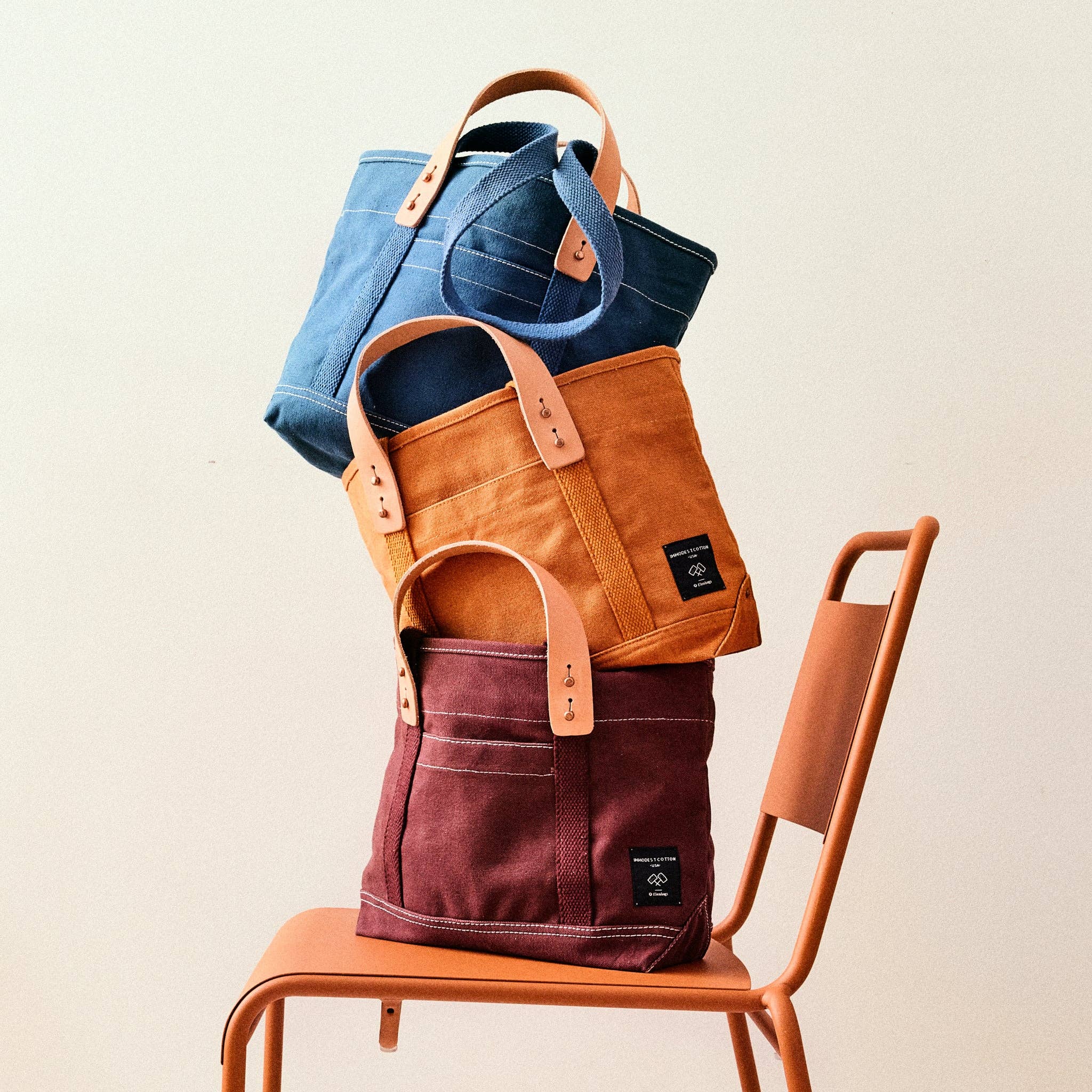 Three canvas tote bags stacked on a rust chair in blue, orange, and maroon colors featuring Lunch Tote | Mustard Seed