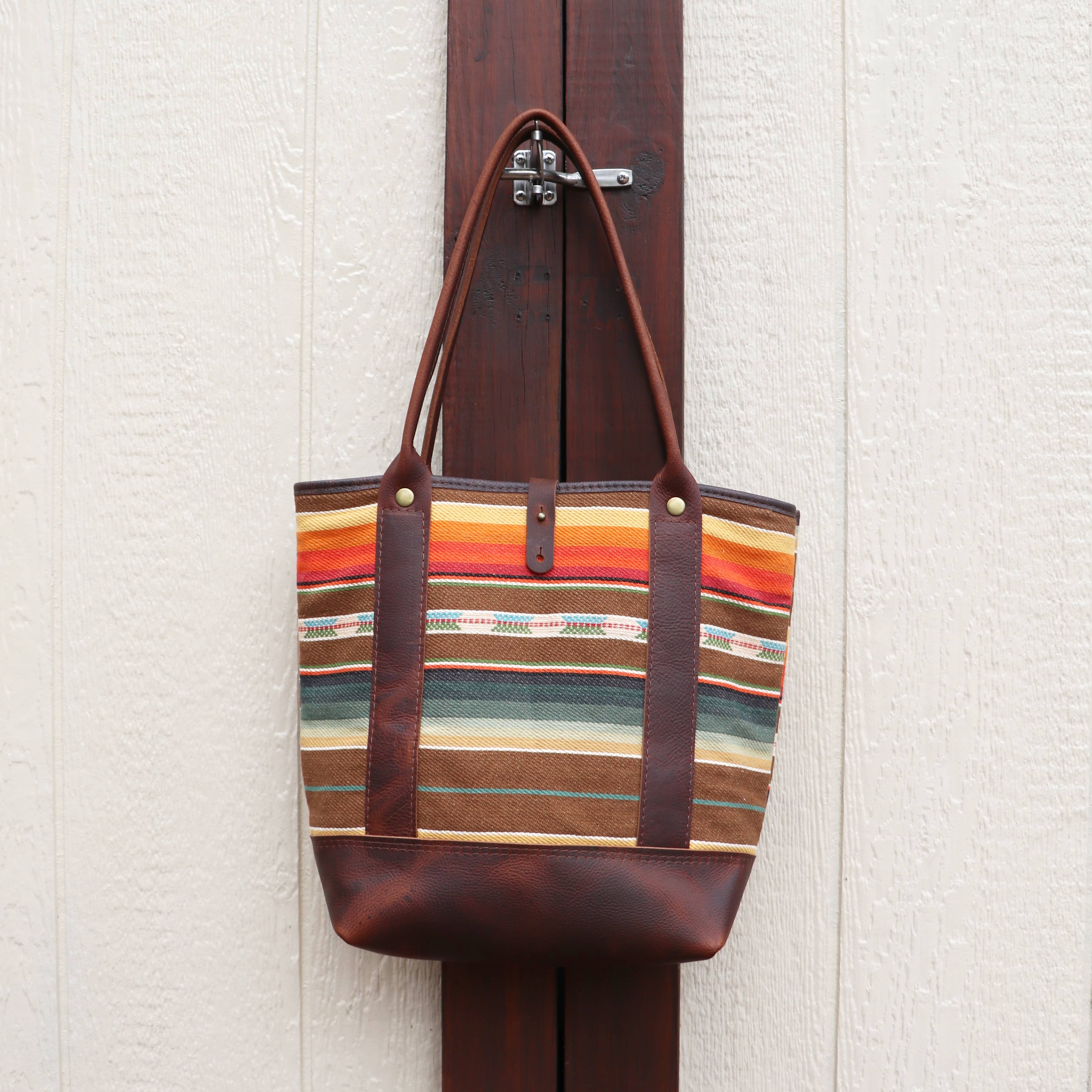 The Pearl Serape Tote by Hawks & Doves, featuring colorful serape stripes and brown leather handles, hangs on a dark wooden door latch against a light textured wall.