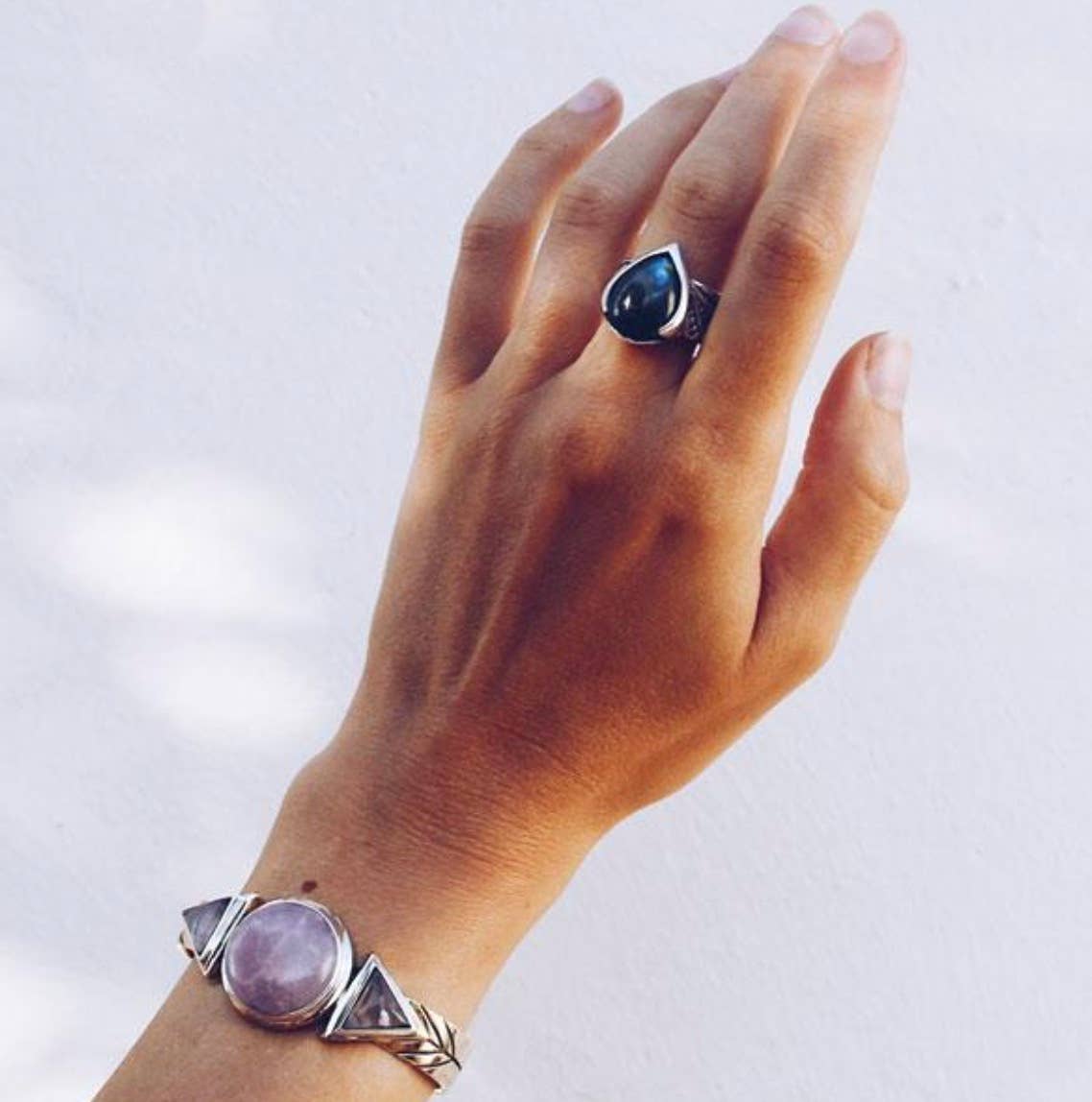 A hand wearing a silver bracelet with a large round purple stone, two triangular stones, and the bahgsu jewels Winged Ring featuring a dark teardrop-shaped stone is shown against a light background.