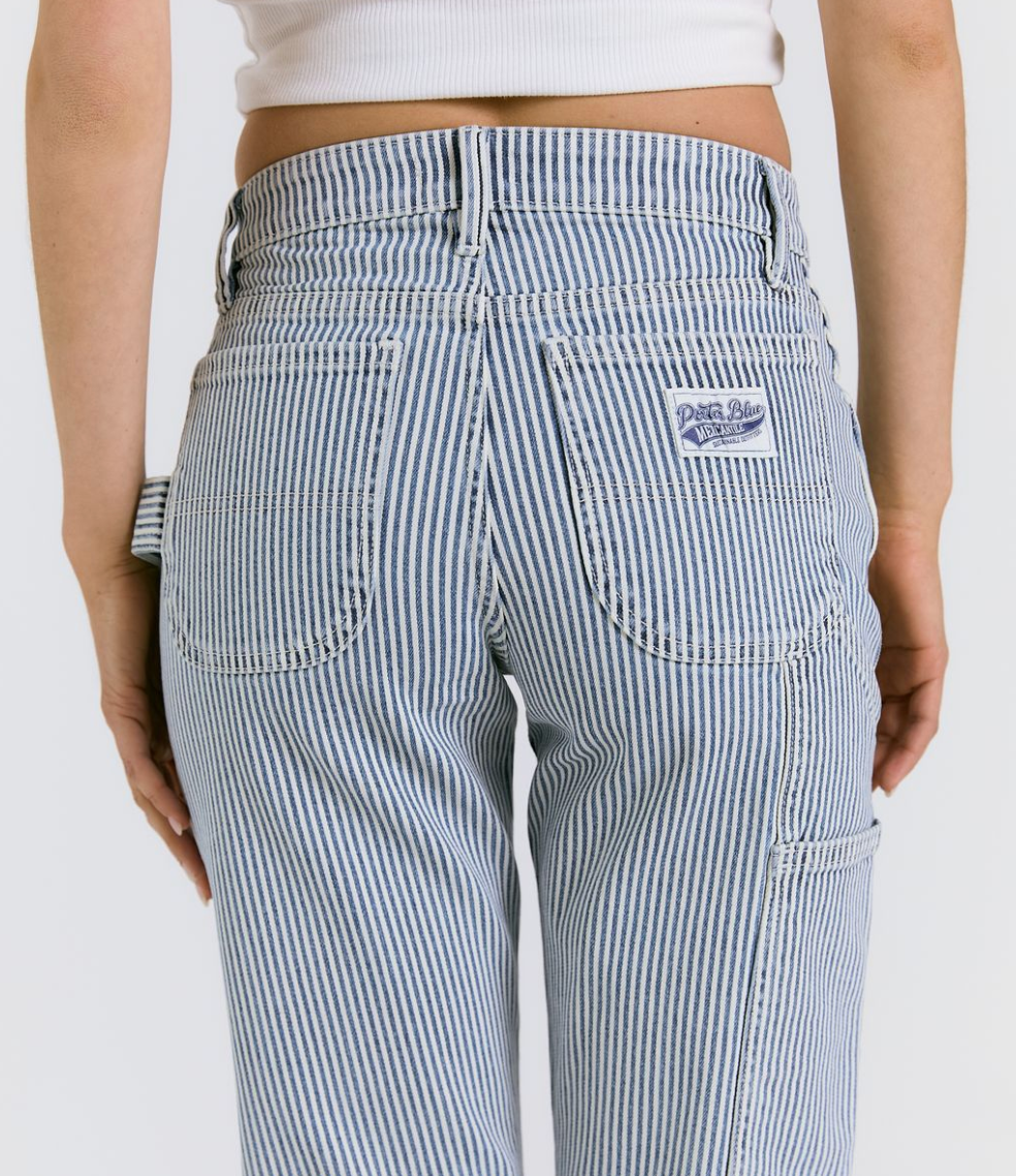 A person is shown from the waist down, facing away, wearing Porter Blue Apparels PEYTON PAINTER IN PACIFIC WASH jeans and a white cropped top. The jeans are vintage-inspired carpenter pants with blue and white stripes.
