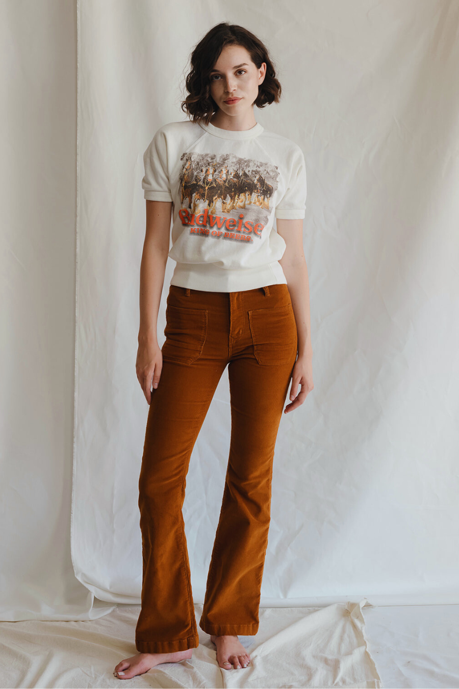 A woman with short dark hair stands barefoot on draped white fabric, wearing the People Of Leisure Budweiser Clydesdale Raglan and high-waisted burnt orange corduroy pants against a plain white backdrop.