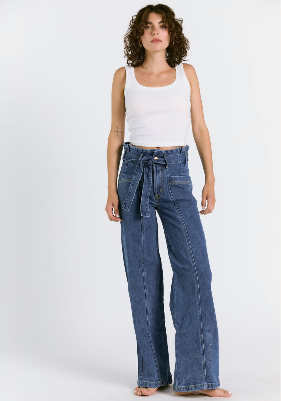 A woman with curly hair models the PENNY PAPERBAG WIDE LEG IN LAGUNA WASH jeans by Porter Blue Apparel, featuring a tie belt and wide-leg silhouette, paired with a white tank top. Made from Candiani Mills organic cotton denim, she stands barefoot against a plain background.