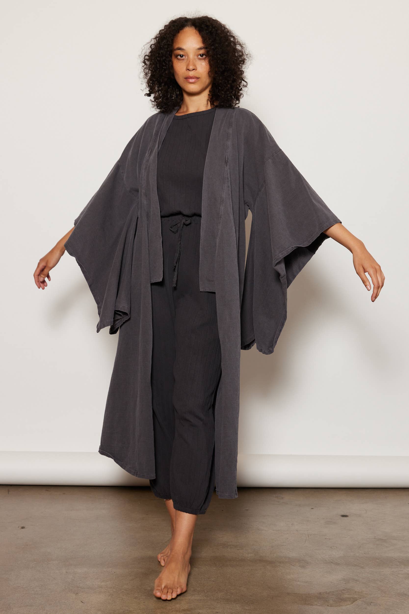 A woman with curly hair poses barefoot against a plain background, wearing the ethically made Dawn Robe by People Of Leisure over a matching black top and pants, her arms extended to highlight the robe’s wide sleeves.