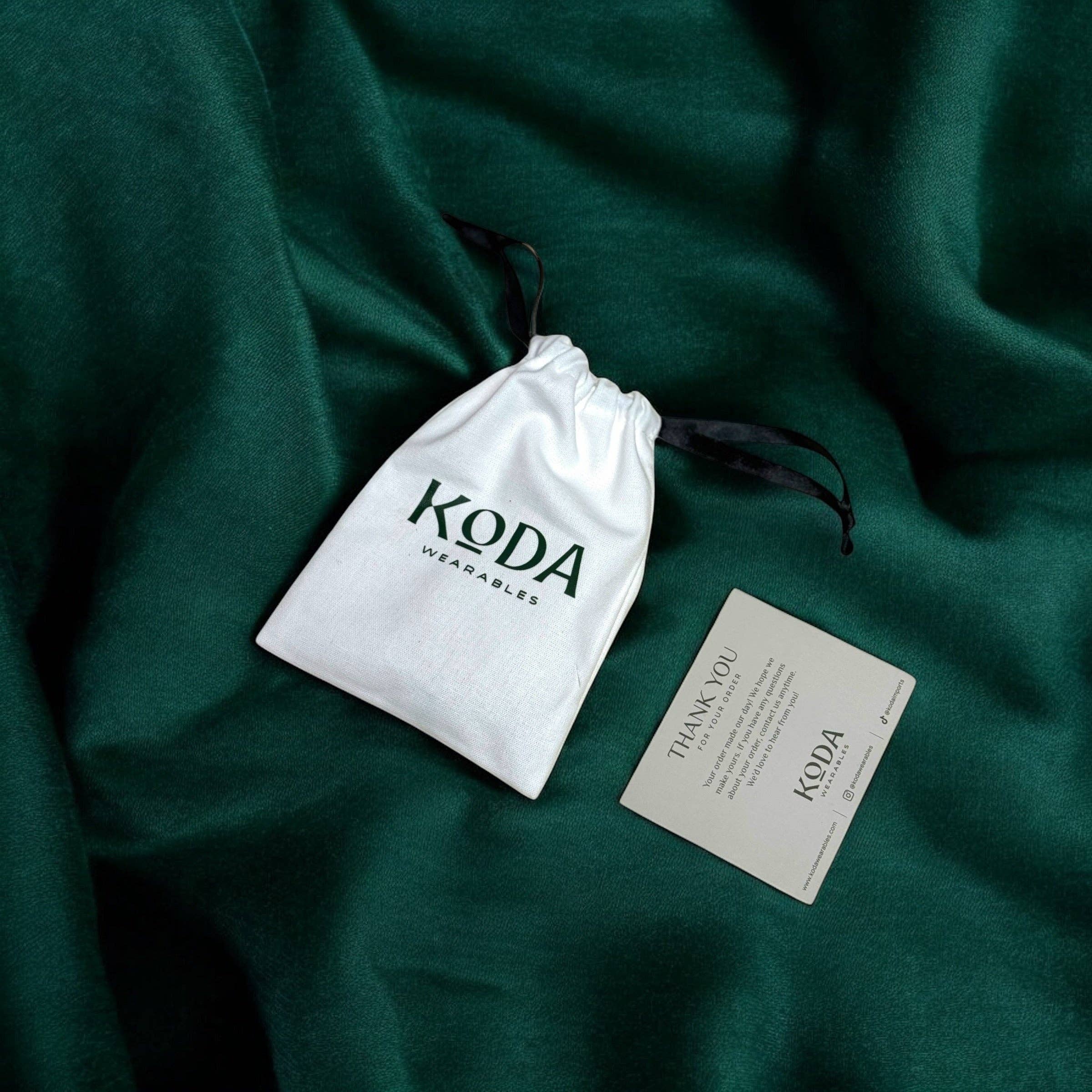 White drawstring pouch and thank you card on green fabric for Cocktail Rings packaging by Koda Wearables