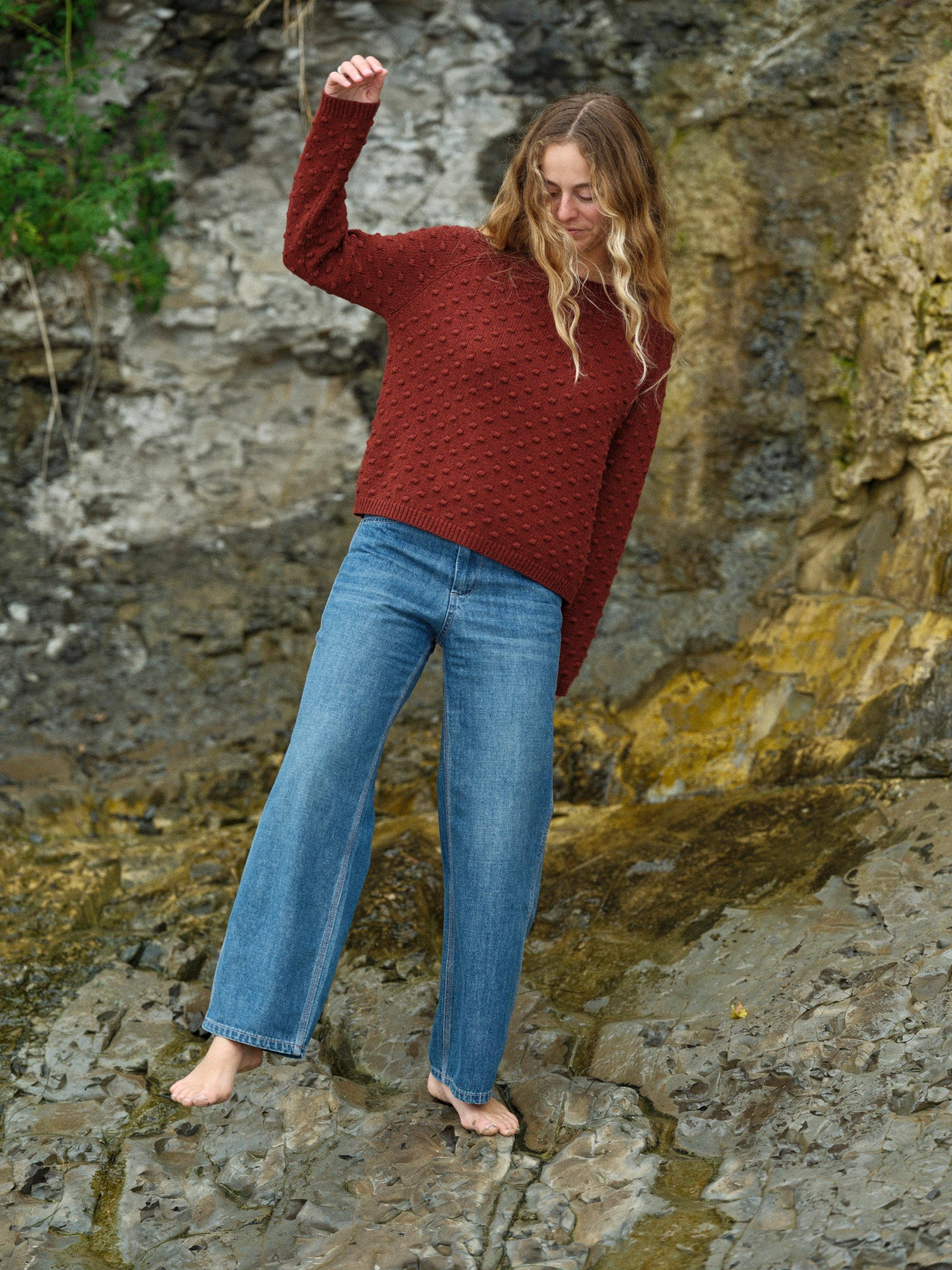 A woman with long wavy hair, wearing the Mollusk Pearl Sweater in red and blue jeans, stands barefoot on rocky terrain before a natural stone wall. She looks down and lifts one arm.