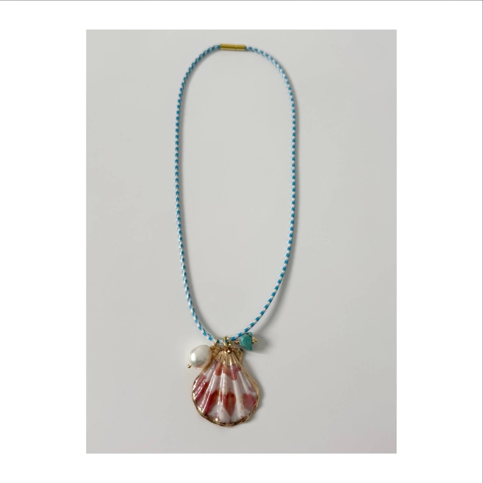 The Islas Necklace by Tramps+Thieves features a blue braided cord, a large pink and white seashell pendant, a freshwater pearl, and a turquoise bead, all showcased on a plain white background.