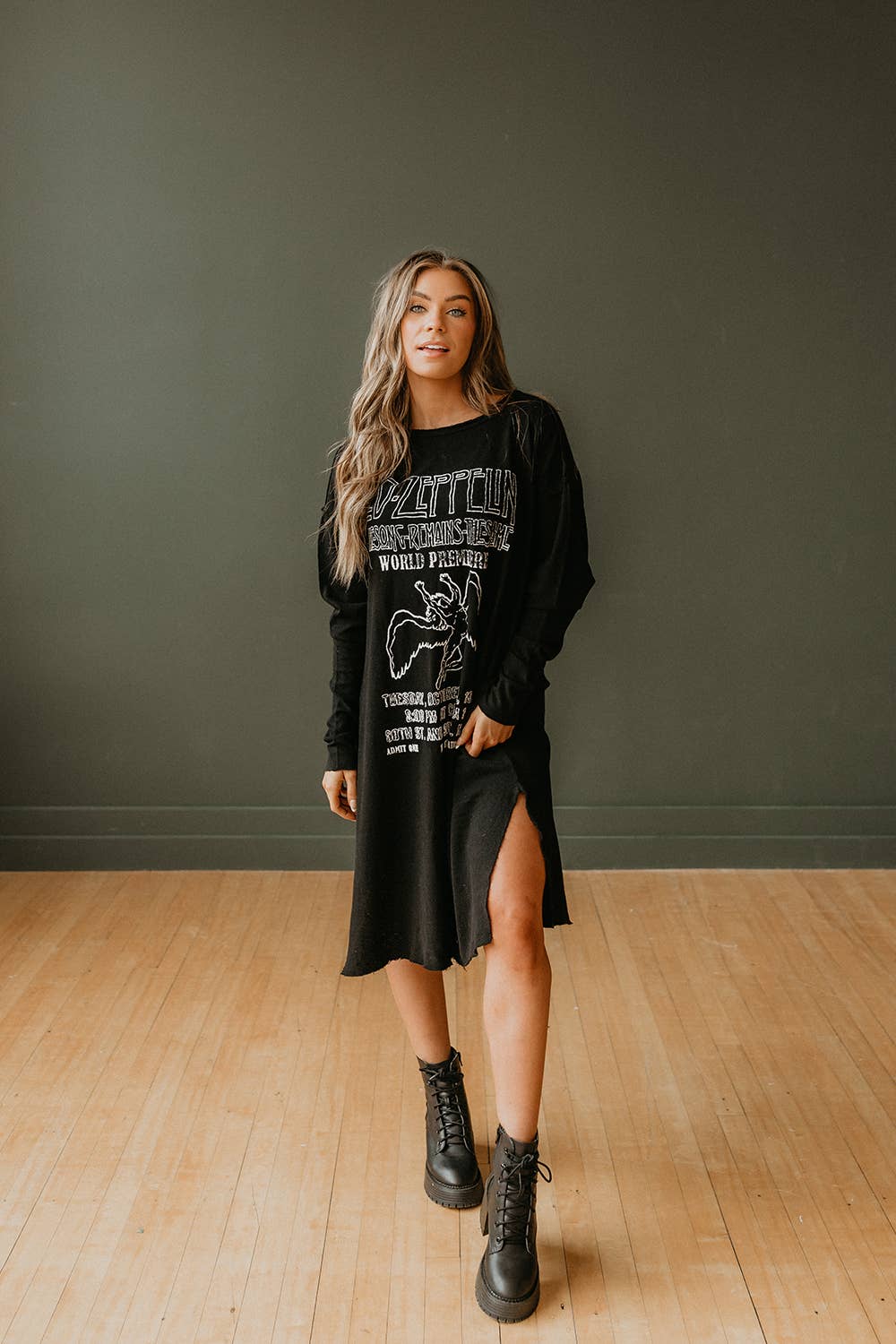 A woman with long wavy hair stands indoors on a wooden floor, confidently modeling the People Of Leisure Led Zeppelin The Song Remains the Same Dress with a thigh-high slit and black lace-up boots against a dark wall.