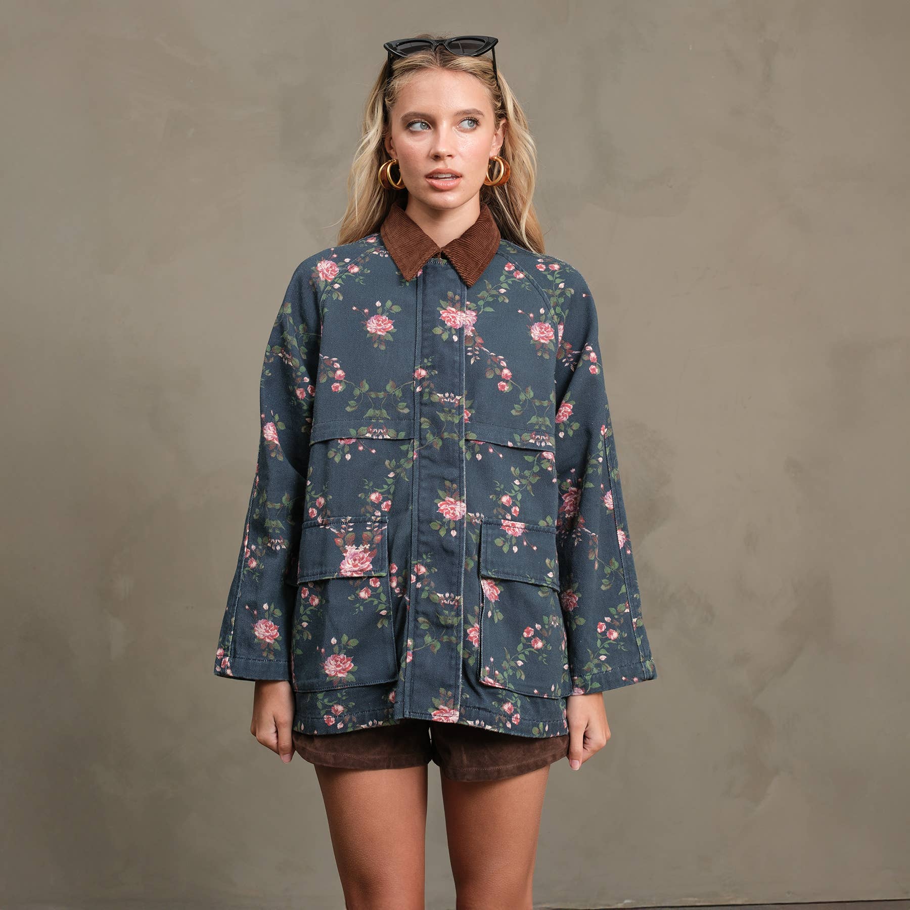 A woman poses against a plain background wearing the RAISONNEL Floral Printed Utility Jacket - RJ1073, featuring large front pockets and a brown collar, paired with brown shorts, gold hoop earrings, and sunglasses on her head.