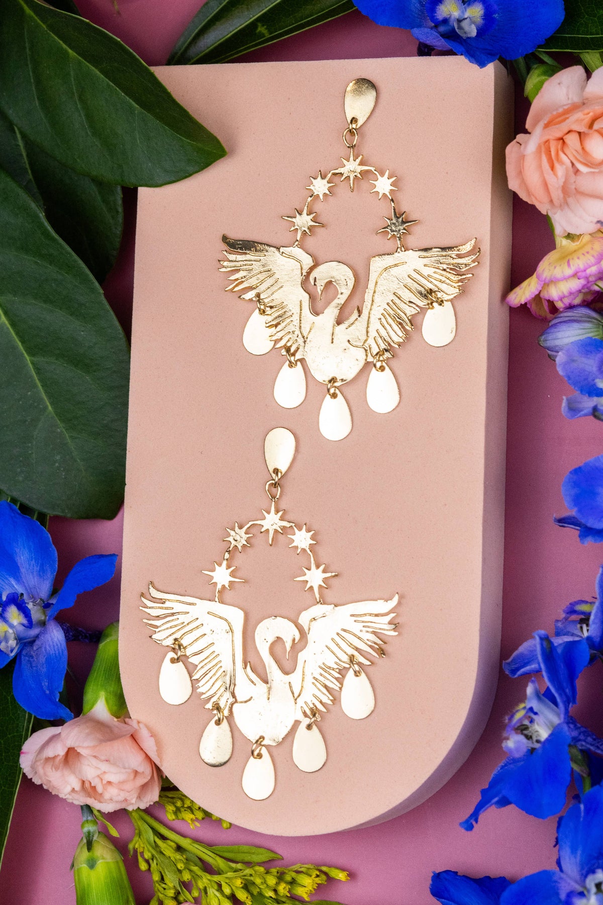 Celestial Swans gold swan earrings with star accents and white teardrop details on a floral display