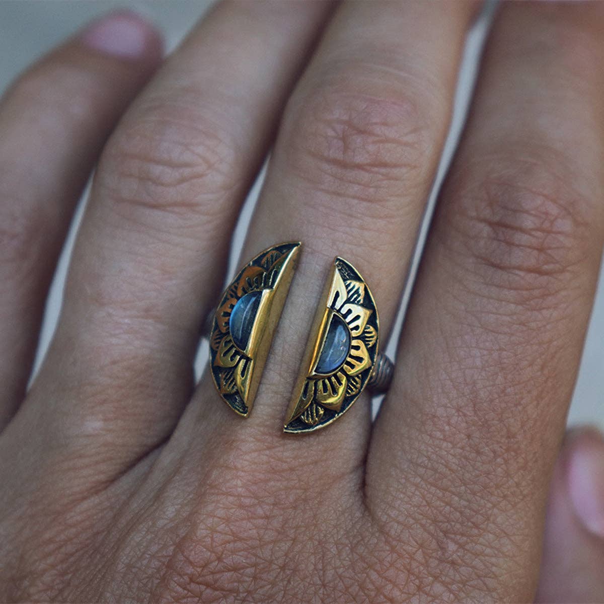 A hand displays the bahgsu jewels Natural Mystic Ring - Moonstone, an adjustable gold and black split-circle design with sun motifs, worn on the middle finger.