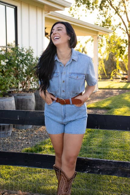 A woman in a denim romper, Ginger Blue Classic Wide Braided Leather Belt in Cognac, and cowboy boots stands smiling by a black wooden fence outdoors, with a house and greenery in the background.