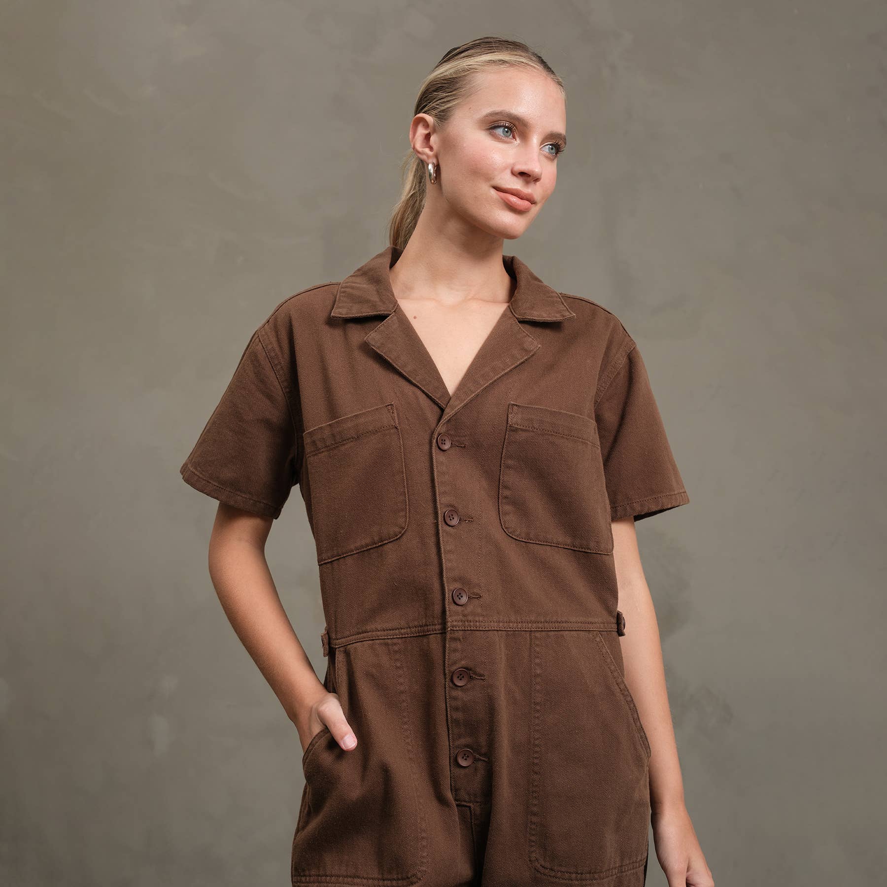 A woman with blond hair pulled back models the RAISONNEL Field Jumpsuit (RJP1001), a brown cotton utility button-down, standing against a gray background with one hand in her pocket, looking slightly to the side and smiling softly.