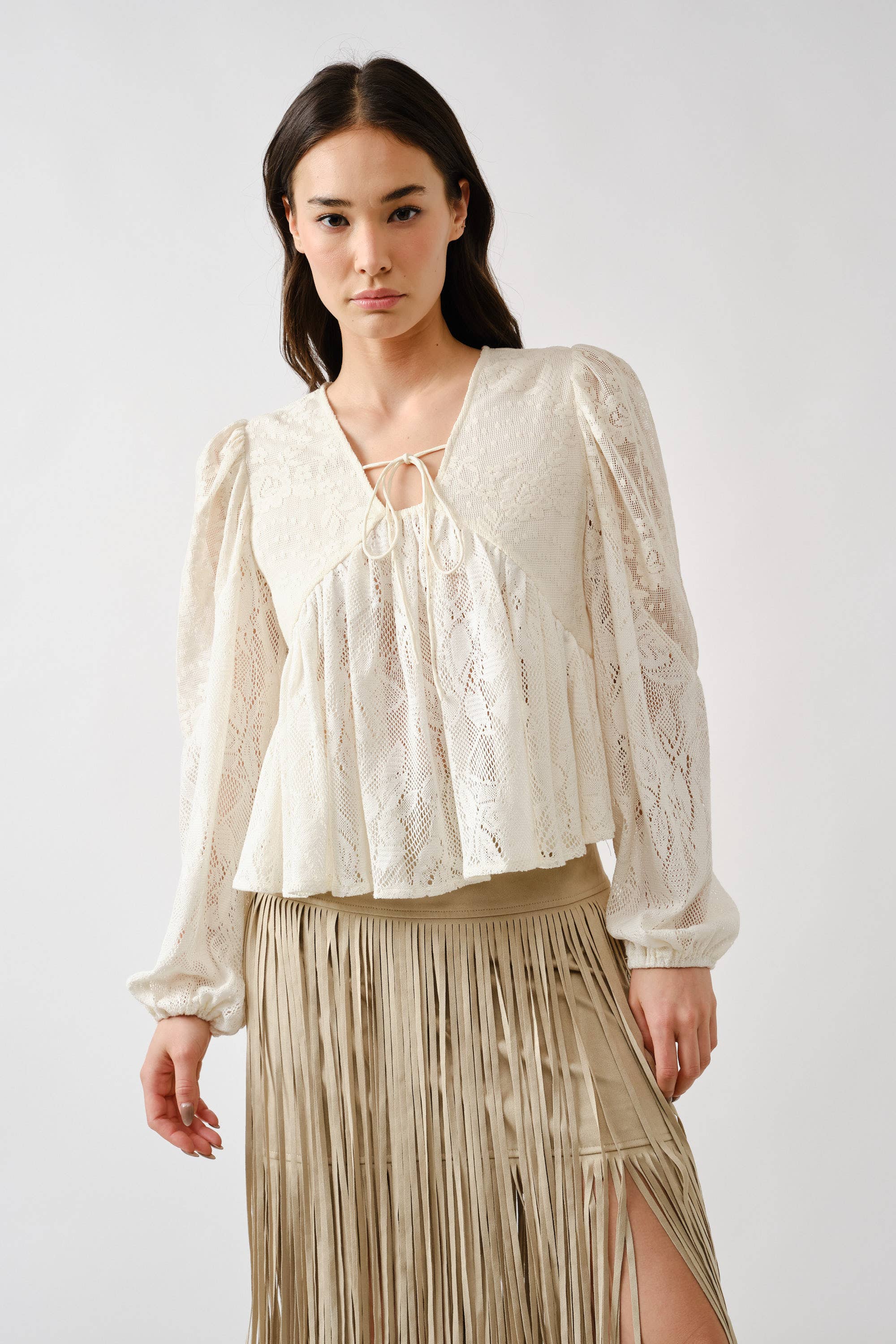 A woman wears the Aaron & Amber Long Sleeve Lace Babydoll, a cream floral lace top with neckline tie, paired with a beige fringe skirt, standing against a white background.