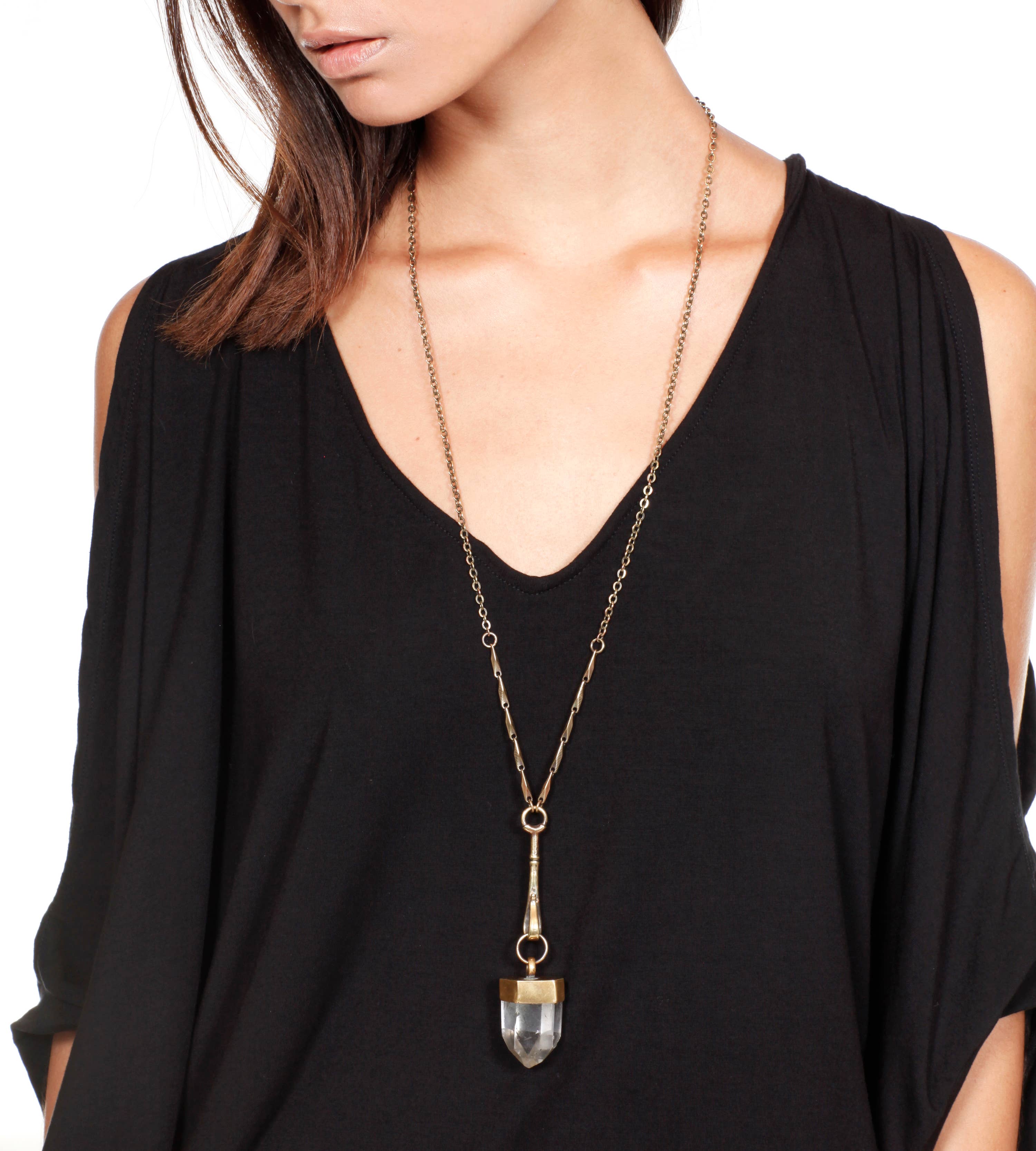A woman wears a loose black top and the Jan Hilmer + Sparrow Crystal Pendulum Necklace Brass, featuring a striking quartz crystal pendant, set against a plain white background.