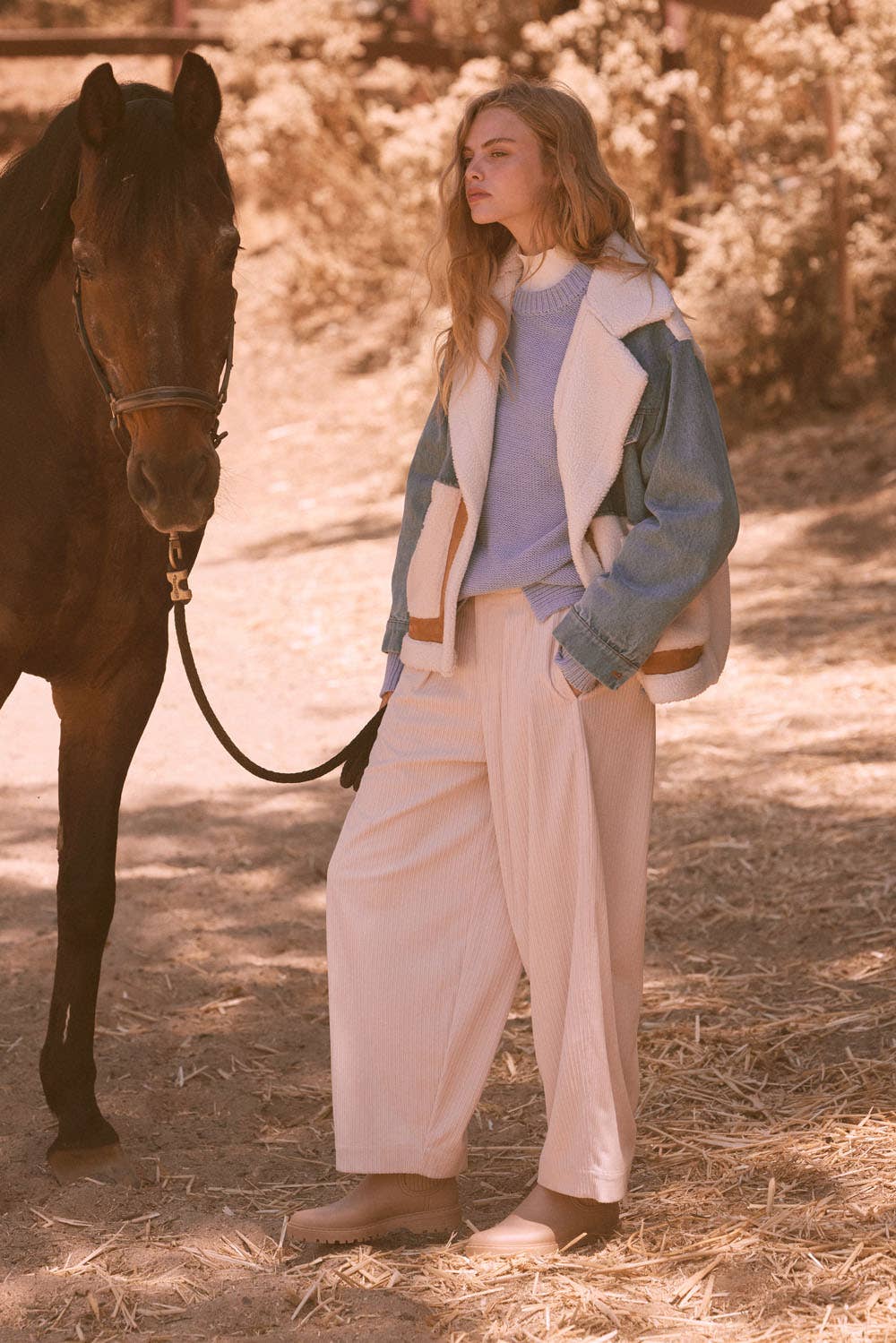 A young woman stands outdoors beside a dark horse, wearing Promesa USA Velvet Corduroy Pleat-Front Wide-Leg Pants in cream, a light blue sweater, and a denim jacket with white fleece collar. Straw and trees create a rustic backdrop.