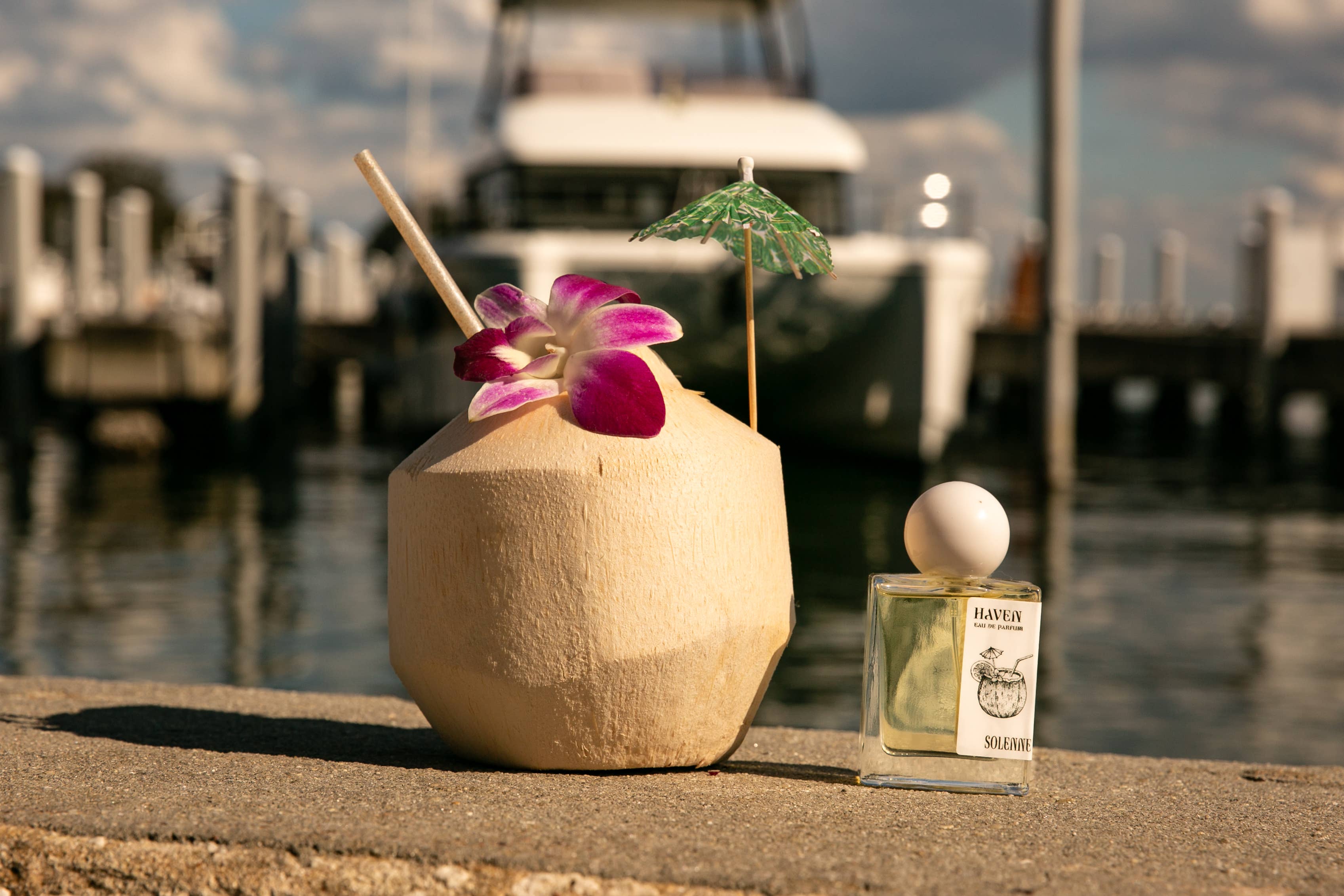 HAVEN - Eau De Parfum - Nontoxic Fragrance bottle by a tropical coconut drink with flower and umbrella near a marina.